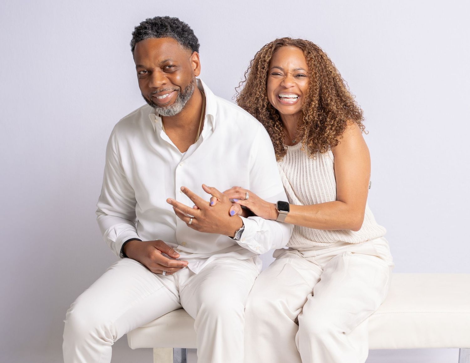 A smiling couple in white outfits poses together on a white bench against a light gray background.