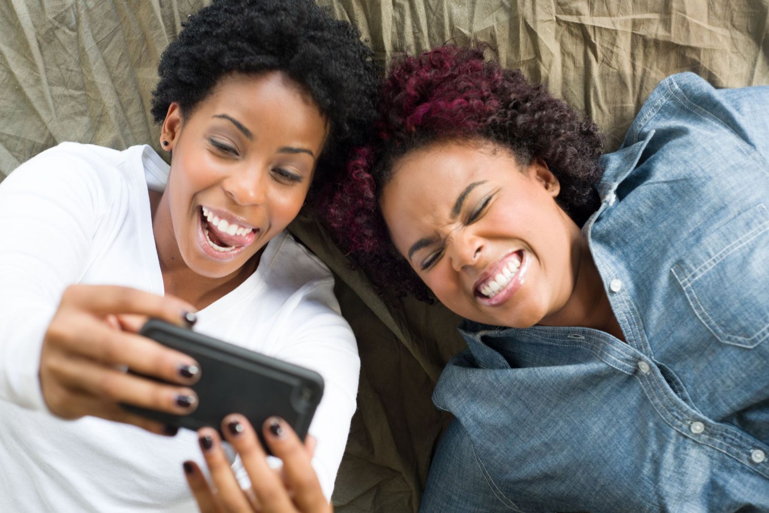 Two friends lying down and laughing while looking at a mobile phone together.