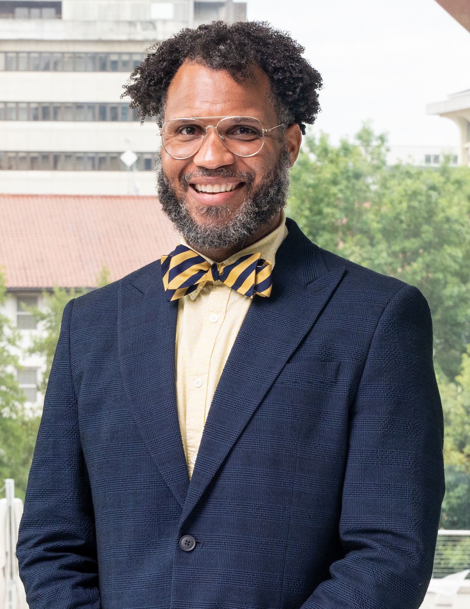 Professional headshot of person wearing a navy suit and gold bowtie smiles warmly in an outdoor business setting.