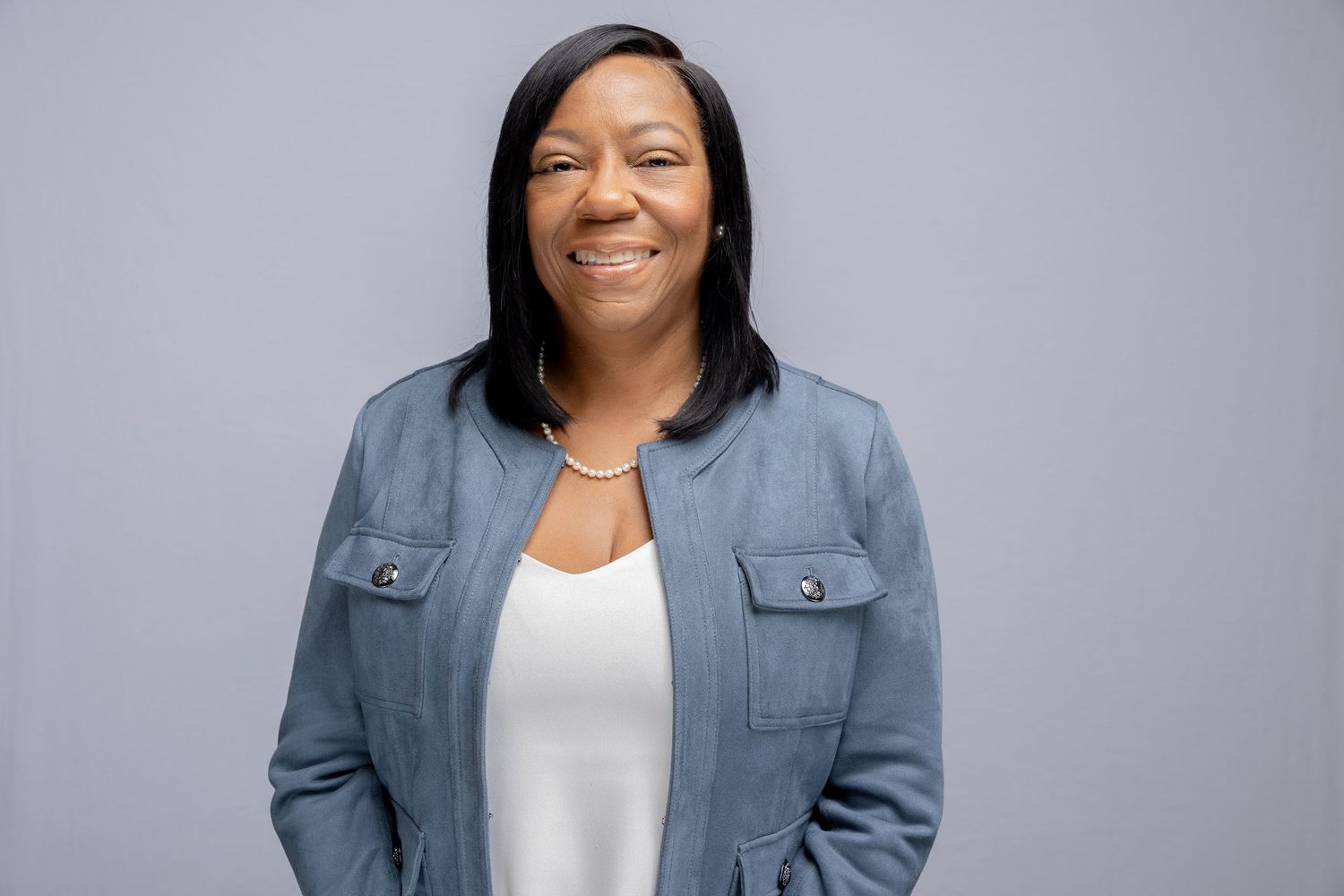 Professional headshot of person wearing a blue denim jacket and white top poses against a gray studio background.