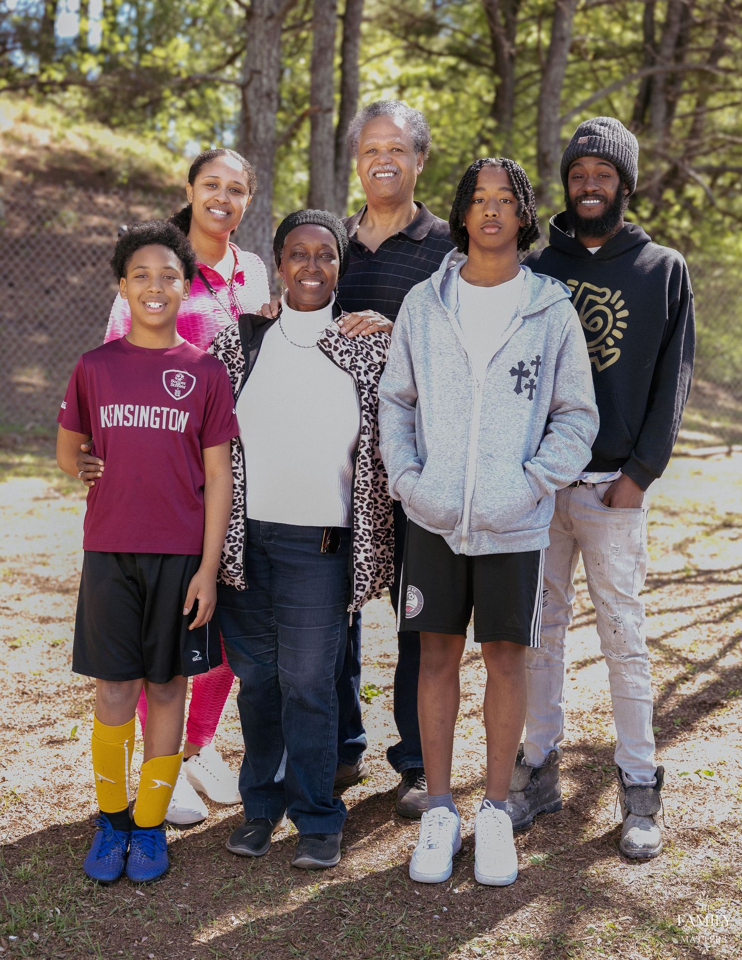 A joyful family of six poses together outdoors among trees on a sunny day.