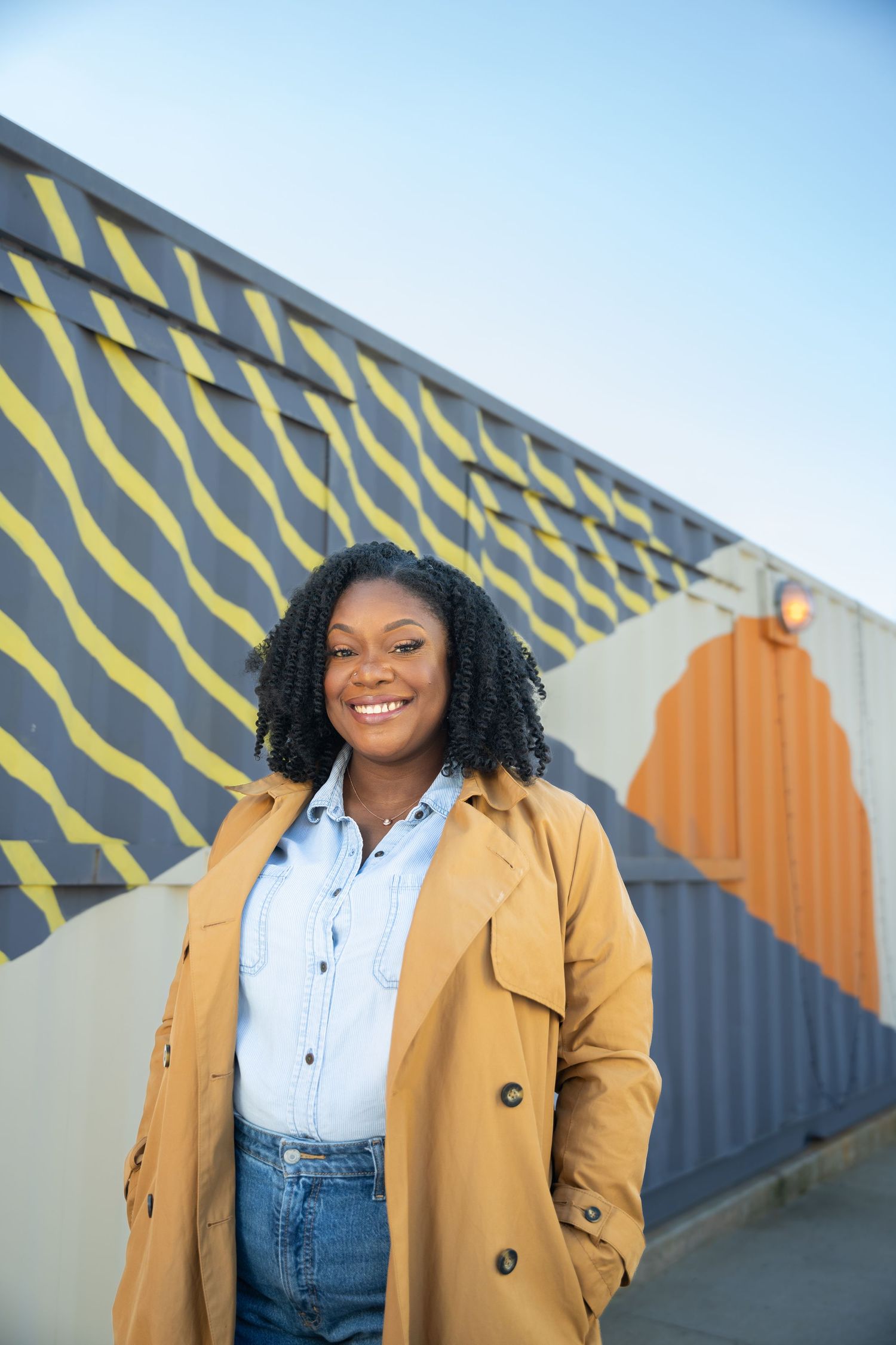 Fashionable outfit featuring a tan trench coat, white button-up shirt and jeans against a geometric painted wall mural.
