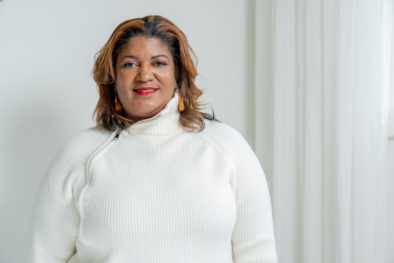 Someone in a white turtleneck sweater smiles warmly against a light-colored background.