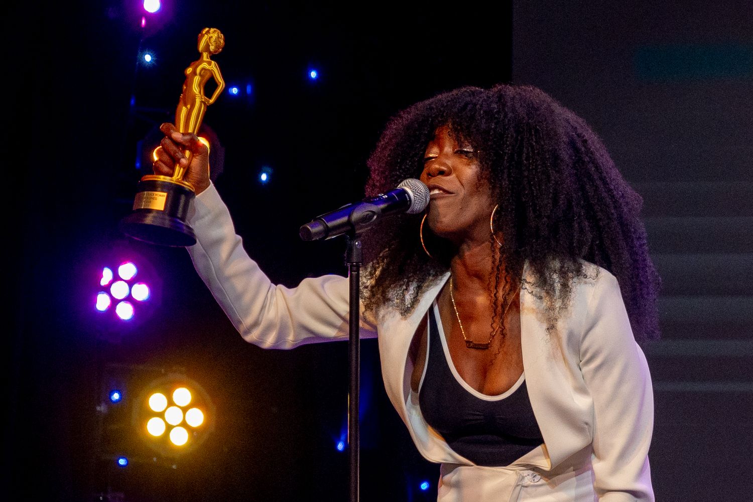 Award recipient in white jacket holds up golden trophy while speaking into microphone with colorful stage lights.