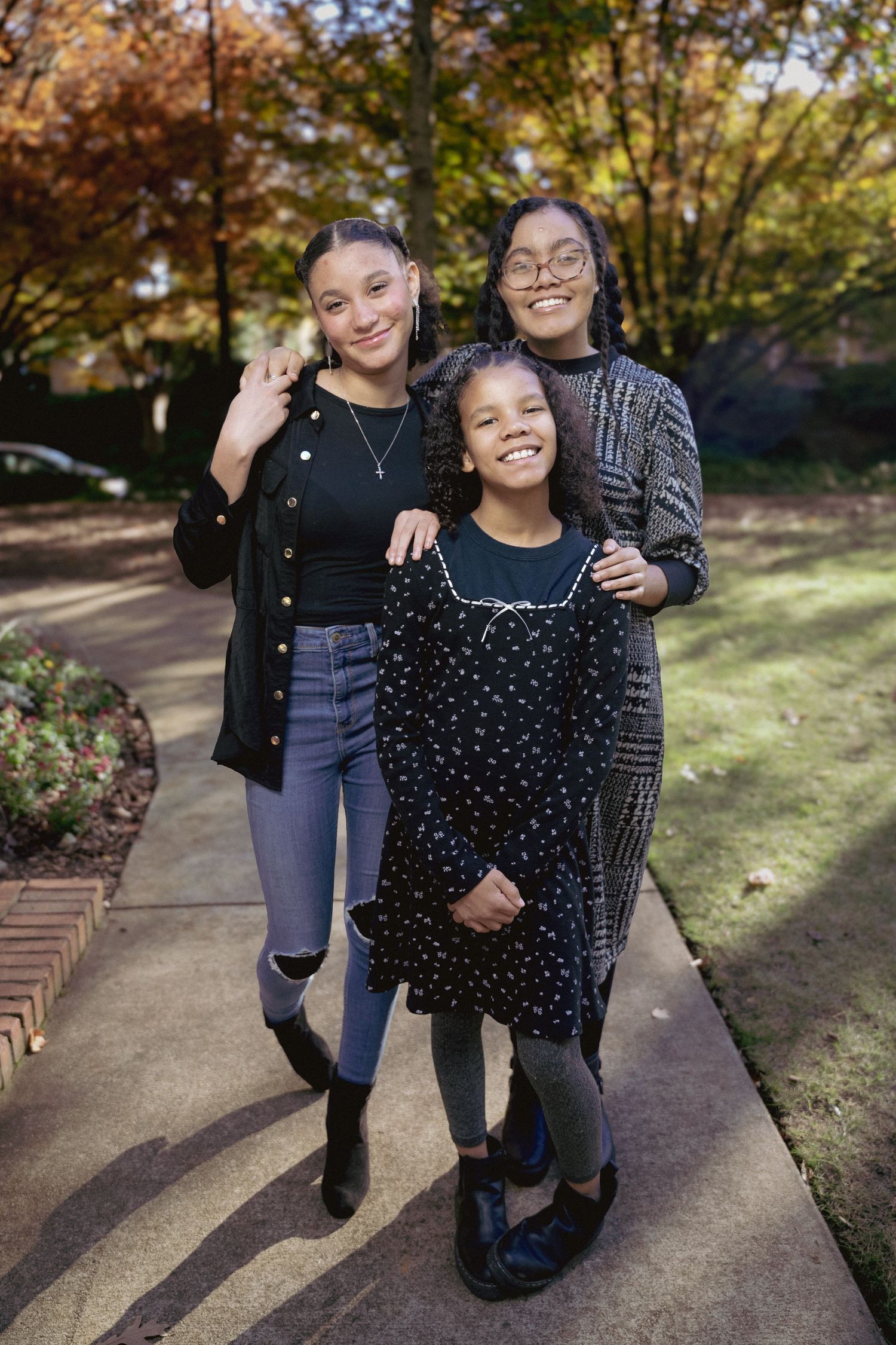 Three smiling girls pose together on a sidewalk surrounded by colorful autumn trees in a park setting.