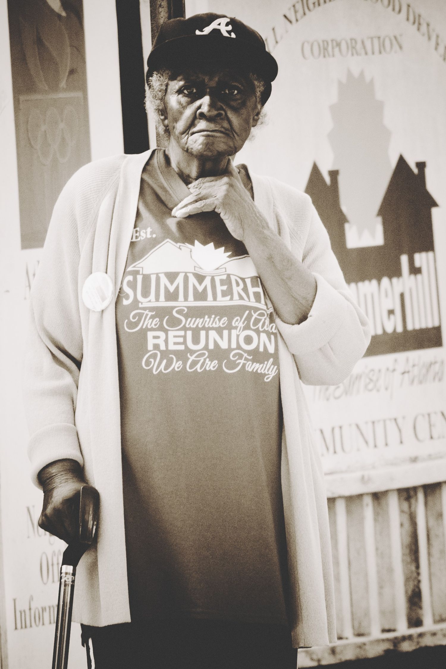 An elderly woman wearing an Atlanta Braves cap and summer reunion t-shirt stands with a cane in a black and white photo.
