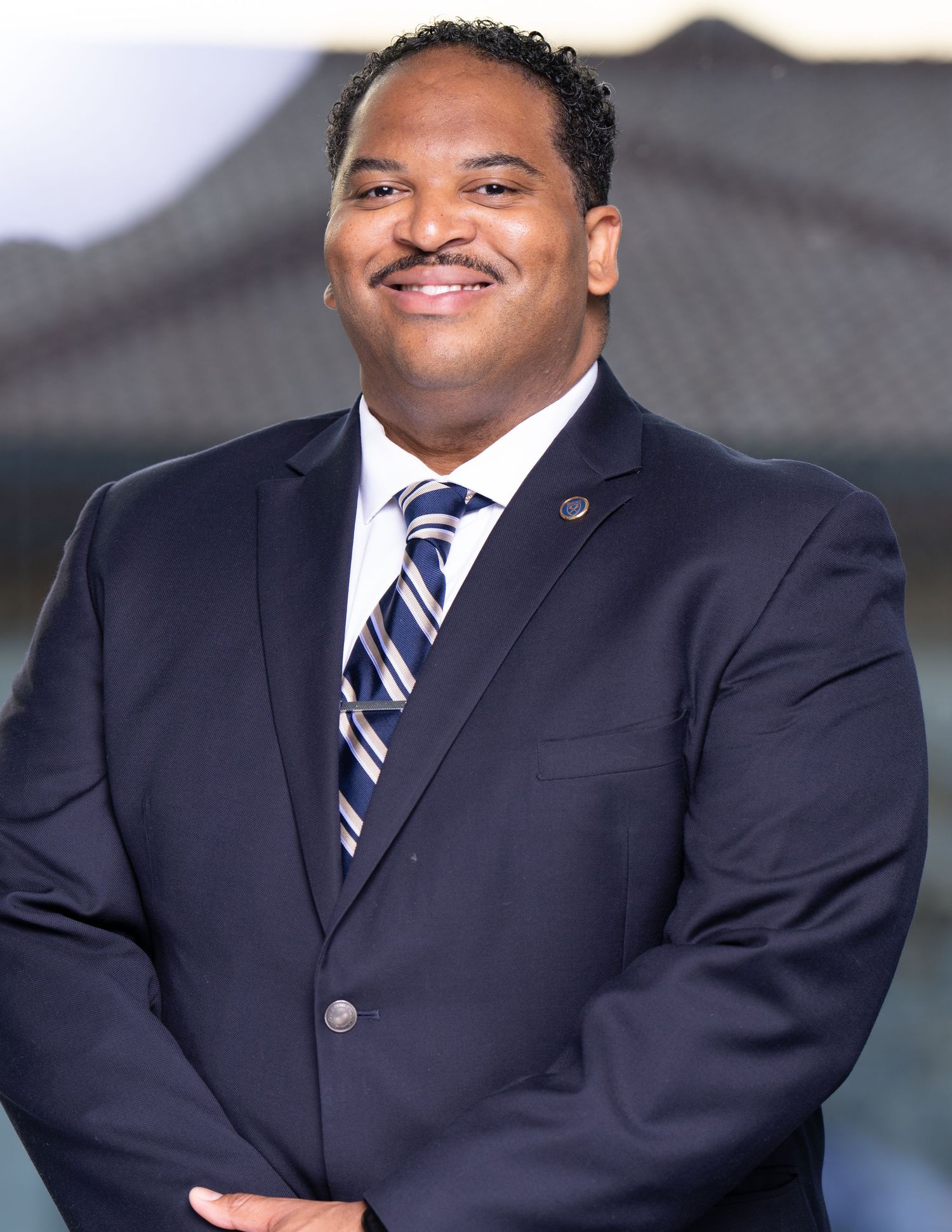 Professional headshot of someone in a navy suit and striped tie smiling warmly against a blurred outdoor background.