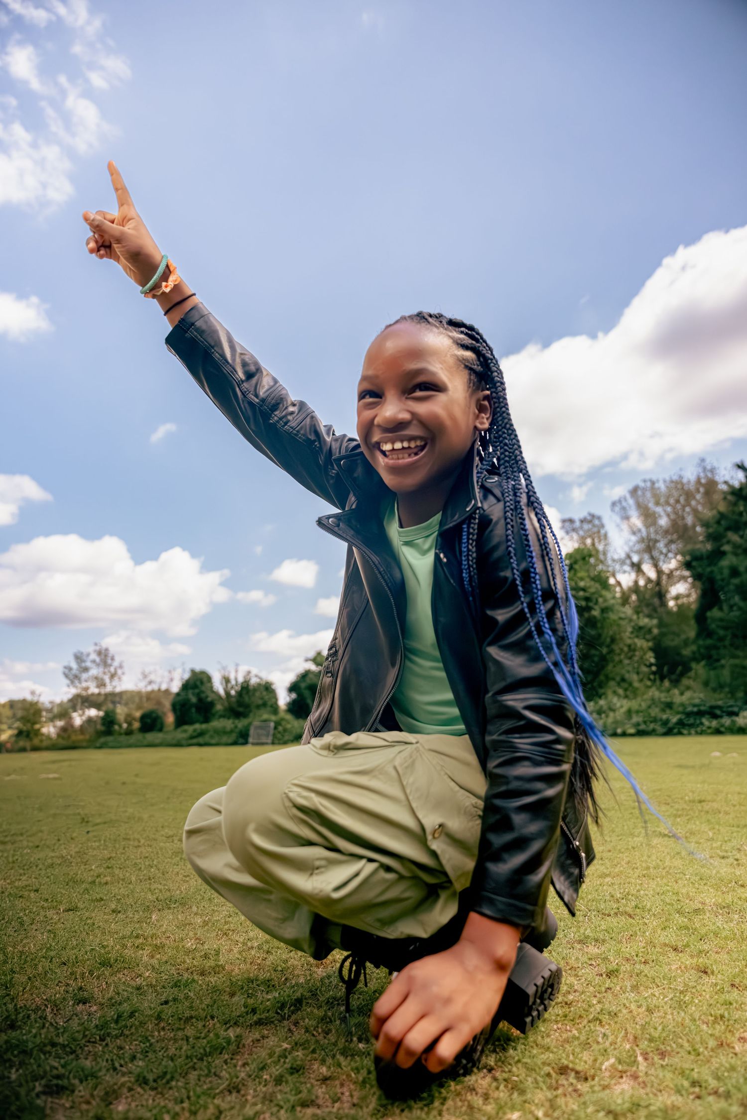 A joyful child wearing a green shirt and black jacket jumps and plays in a grassy field on a sunny day.