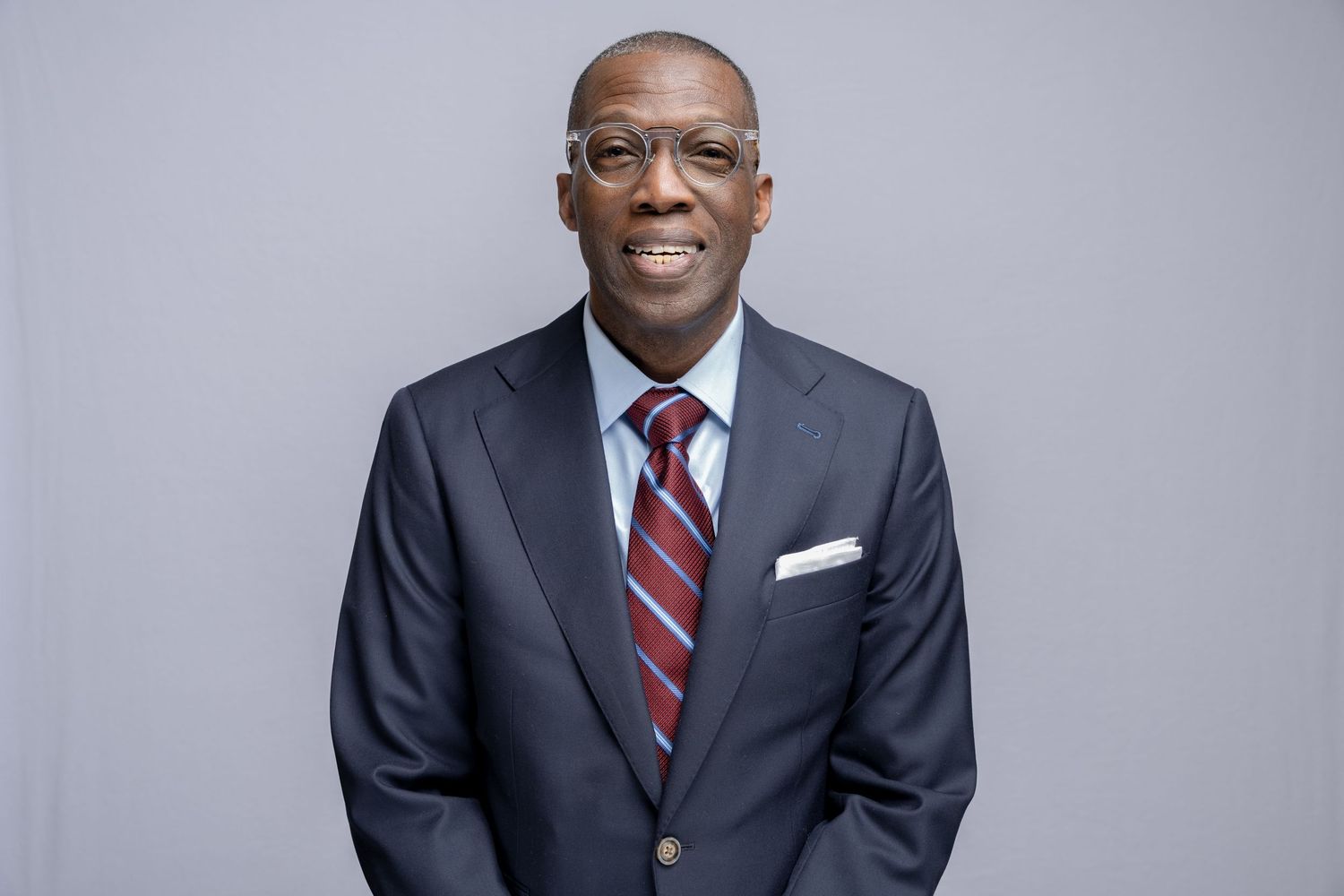 Professional headshot of someone in a dark suit with glasses and red striped tie against a gray background.