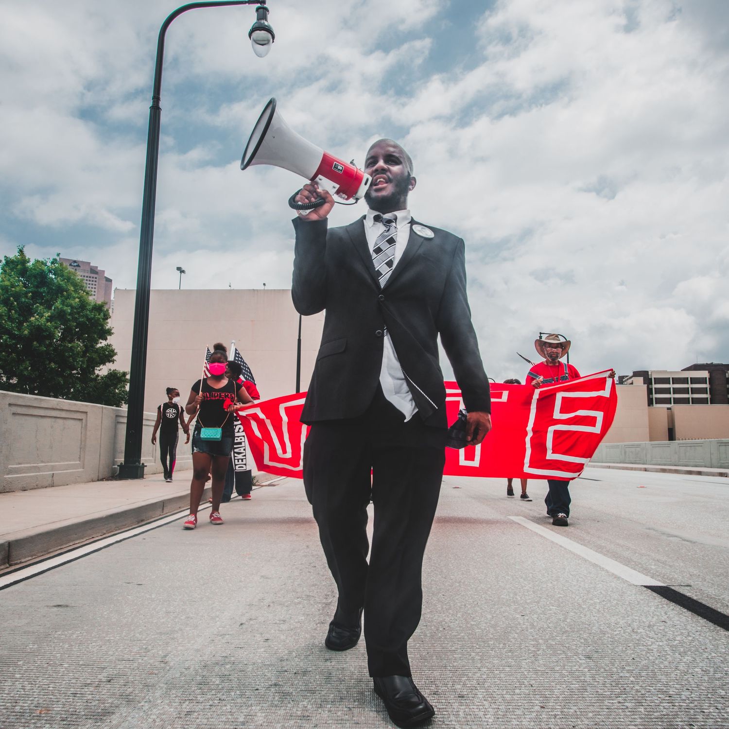 Demonstrator using megaphone during protest march with red signs visible in background.