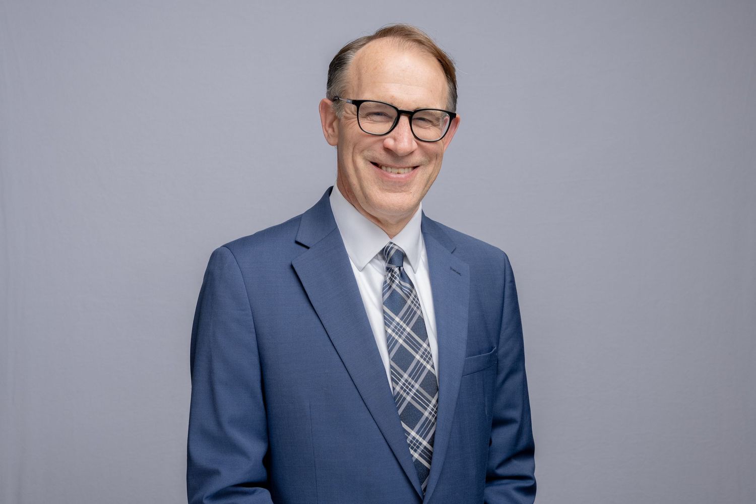 Professional headshot portrait of someone in a blue suit and patterned tie with glasses against a gray background.