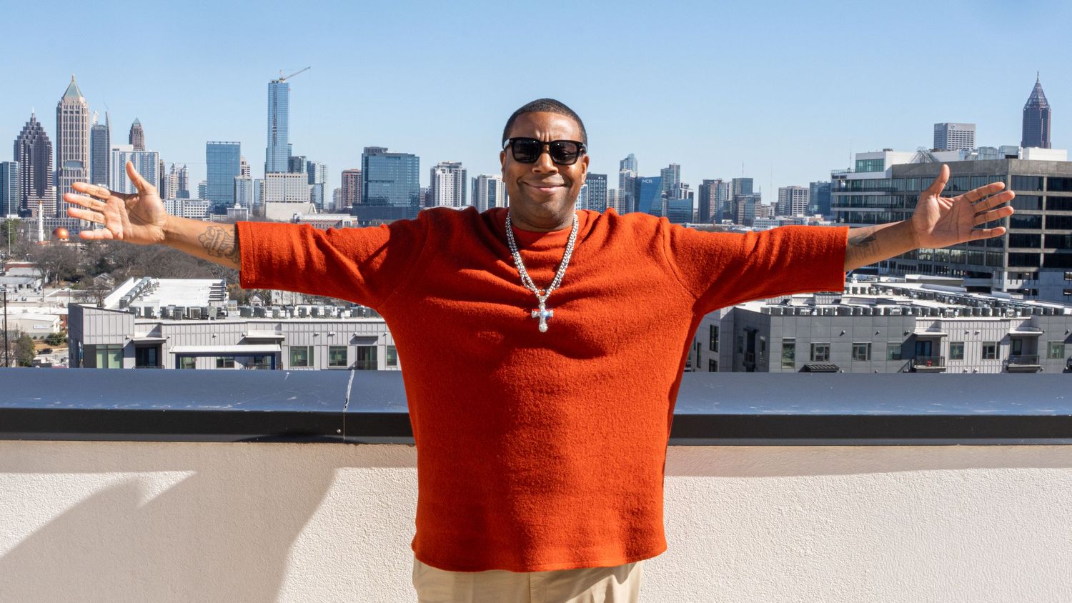 Person in bright orange sweater poses with arms outstretched against Atlanta skyline backdrop on rooftop.