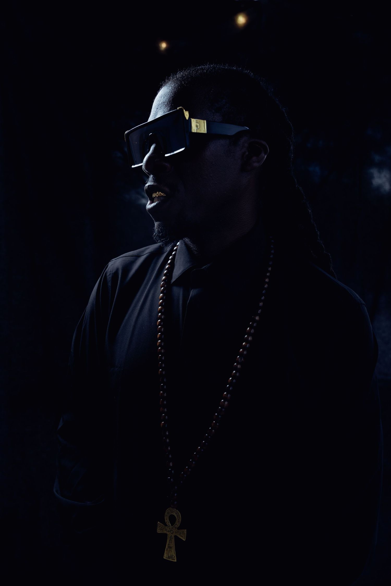 Dramatic low-key portrait of performer wearing sunglasses and ankh necklace against dark background.