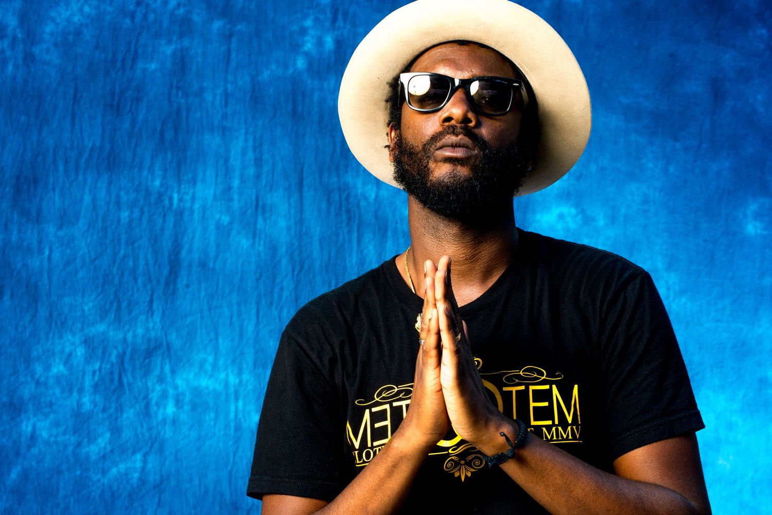 A figure in a white hat and sunglasses poses against a vibrant blue background wearing a black t-shirt.