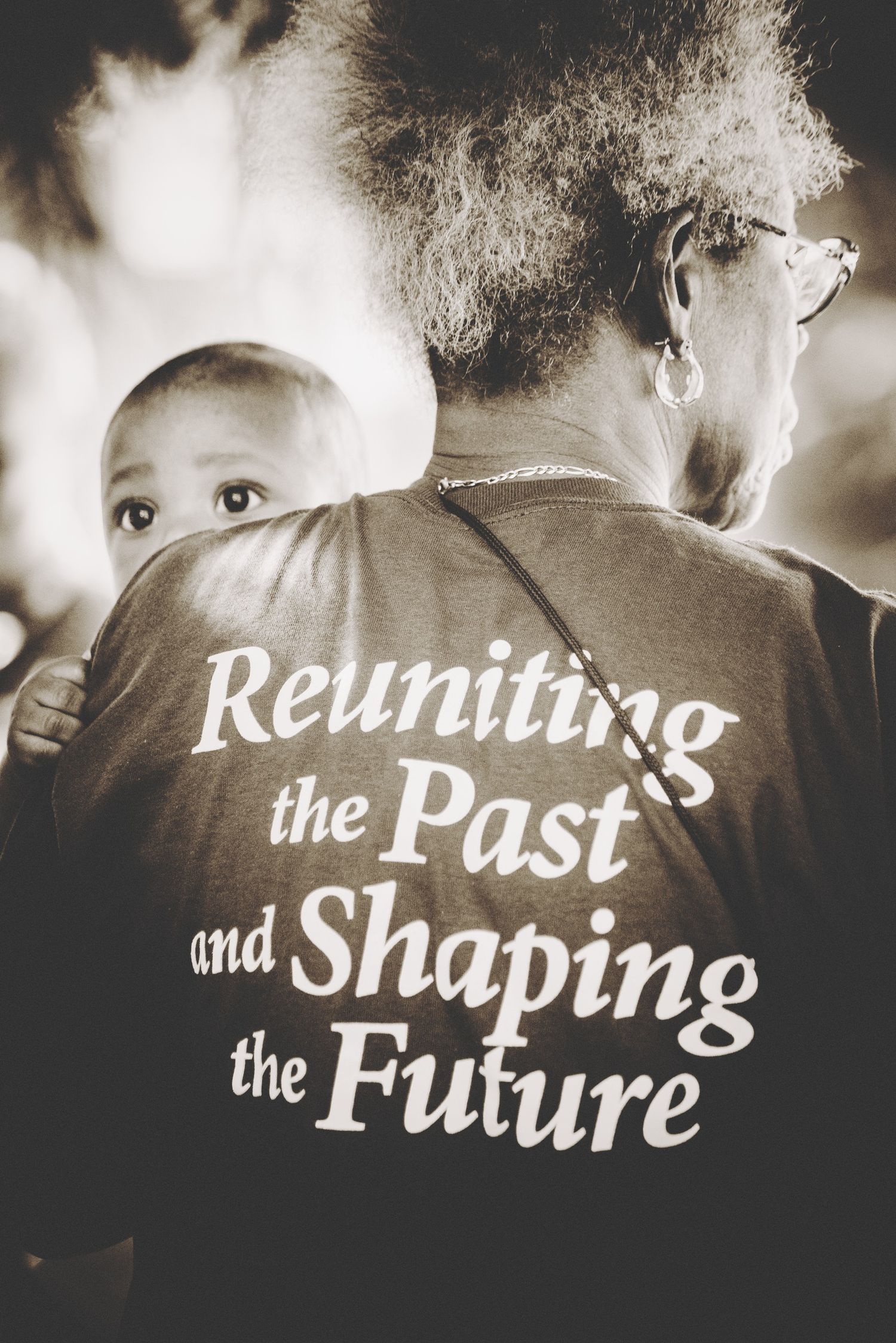 Black and white photo of a baby peeking over an adult's shoulder wearing a shirt reading Reuniting the Past and Shaping the Future.