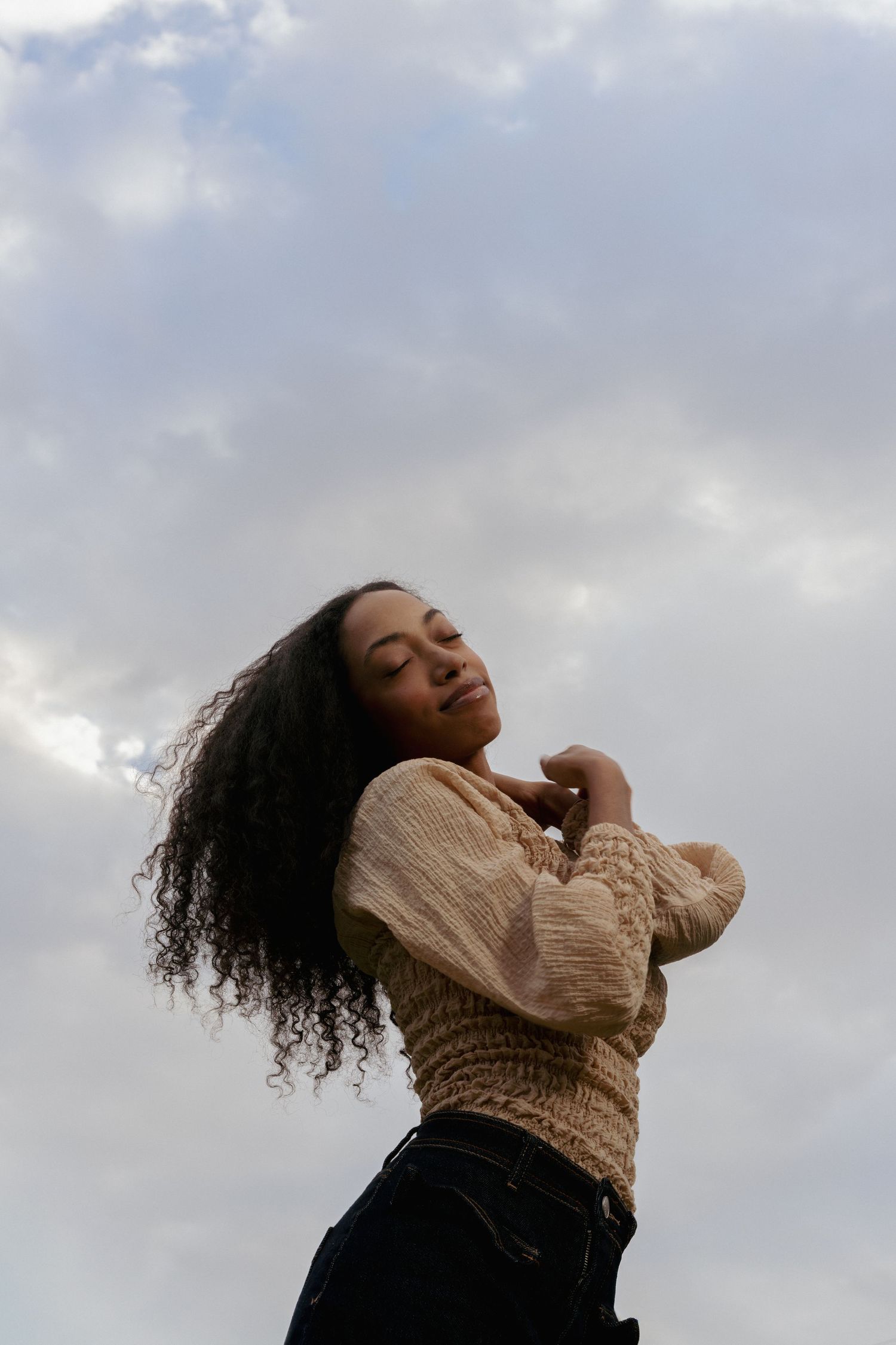 Young woman with curly hair in beige knit sweater posing against cloudy sky, eyes closed, serene expression.