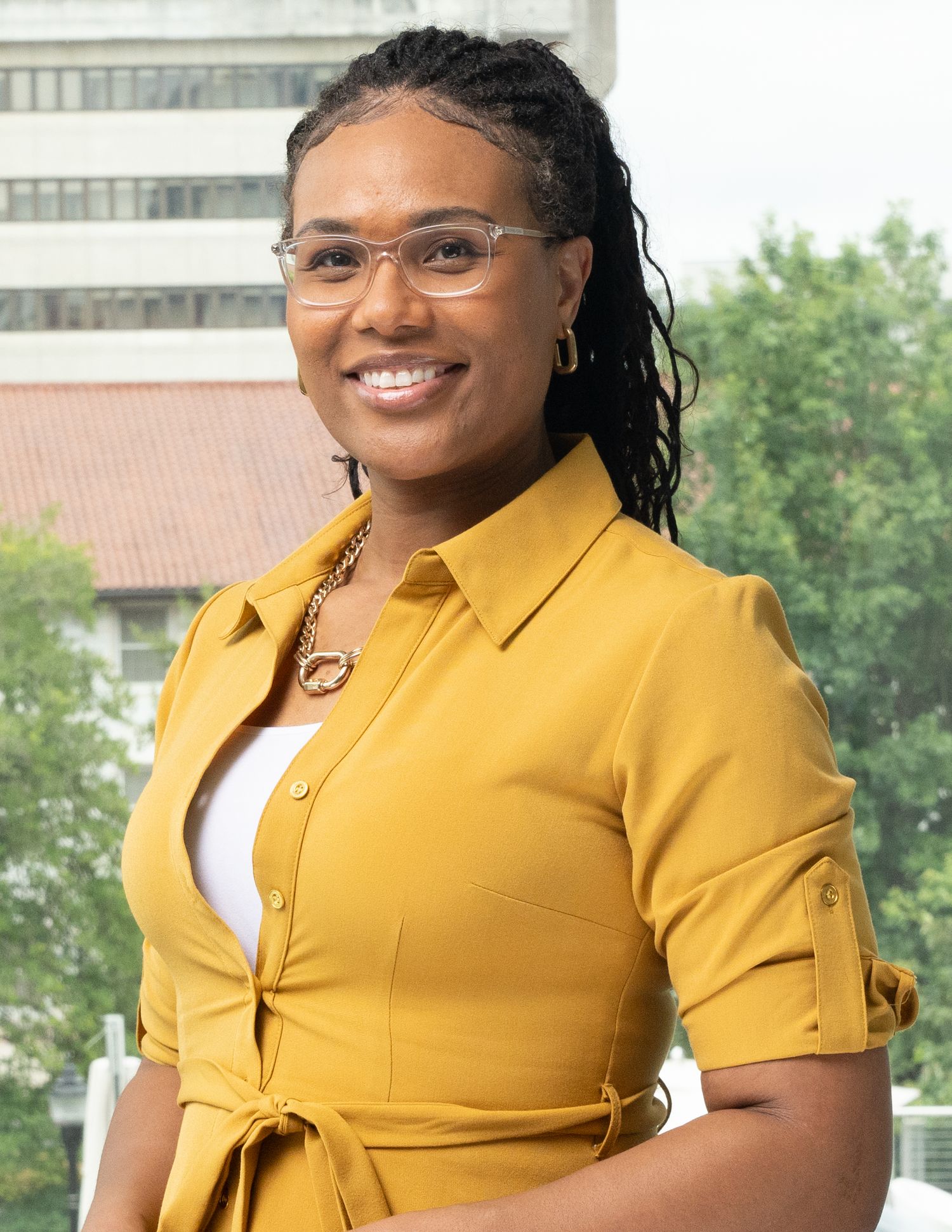 Professional headshot of someone wearing a yellow button-up blouse with glasses and a warm smile outdoors.