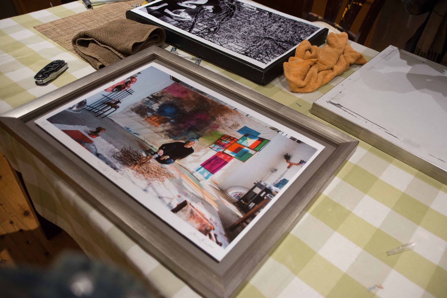 A framed artwork print displayed on a checkered tablecloth surrounded by papers and art supplies in a studio setting.