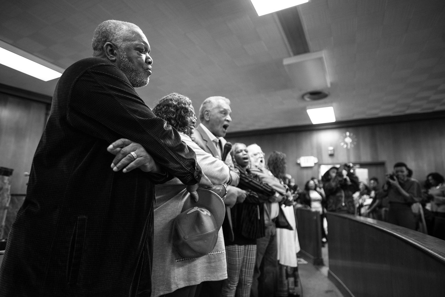 Black and white image showing people singing in what appears to be an indoor meeting or event space.