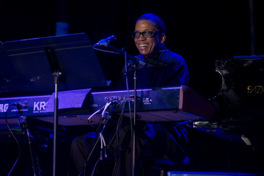 A drummer performs on stage with blue lighting and musical equipment visible in the dark concert setting.