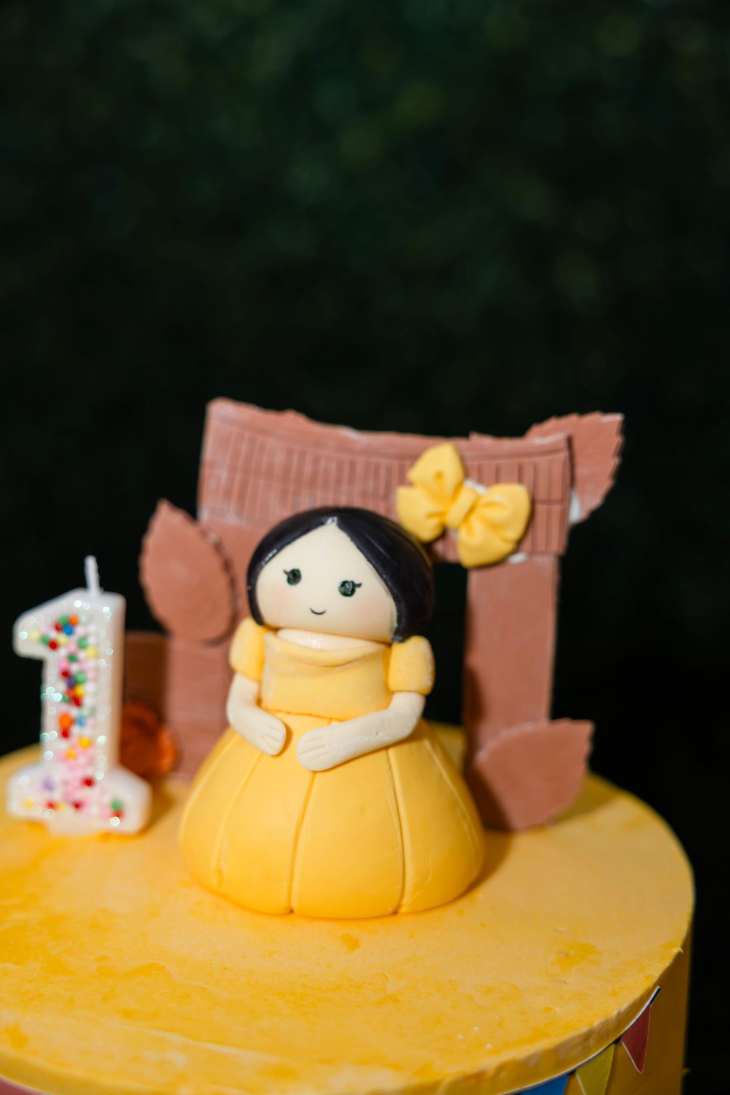 A yellow fondant cake topper of a doll figure with a bow sits next to a number one candle on a cake.