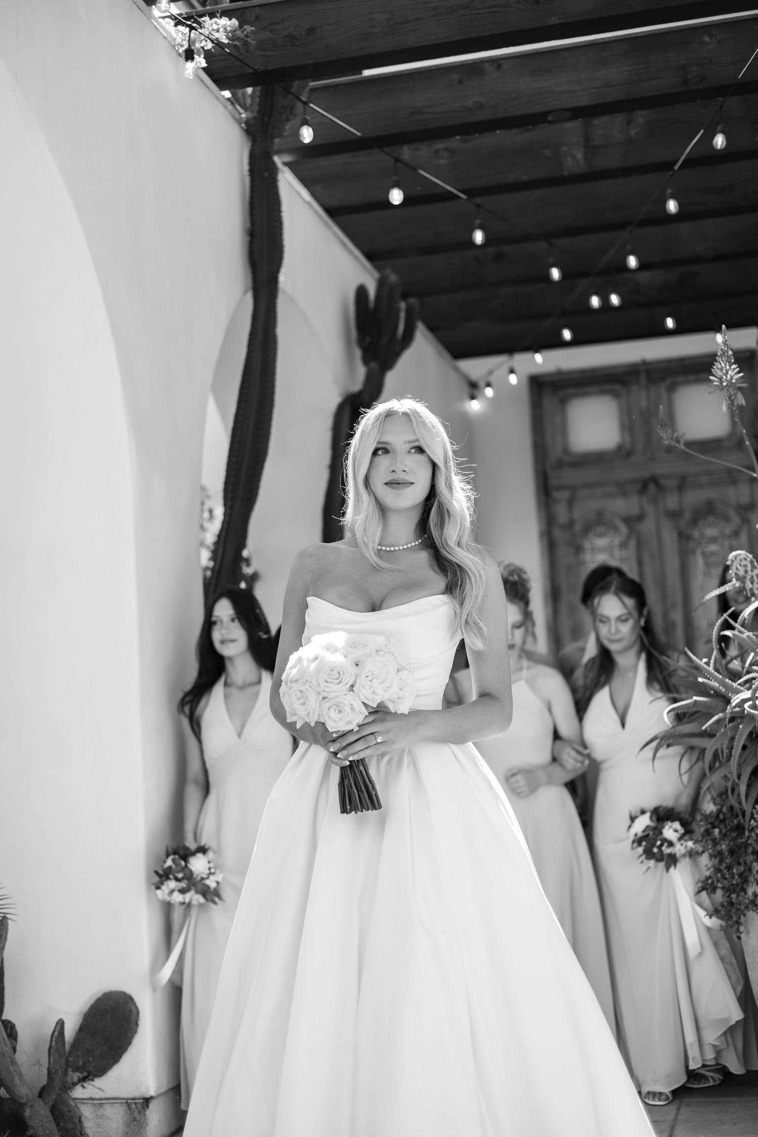 Bridesmaids in elegant white dresses pose together holding bouquets at a wedding celebration.
