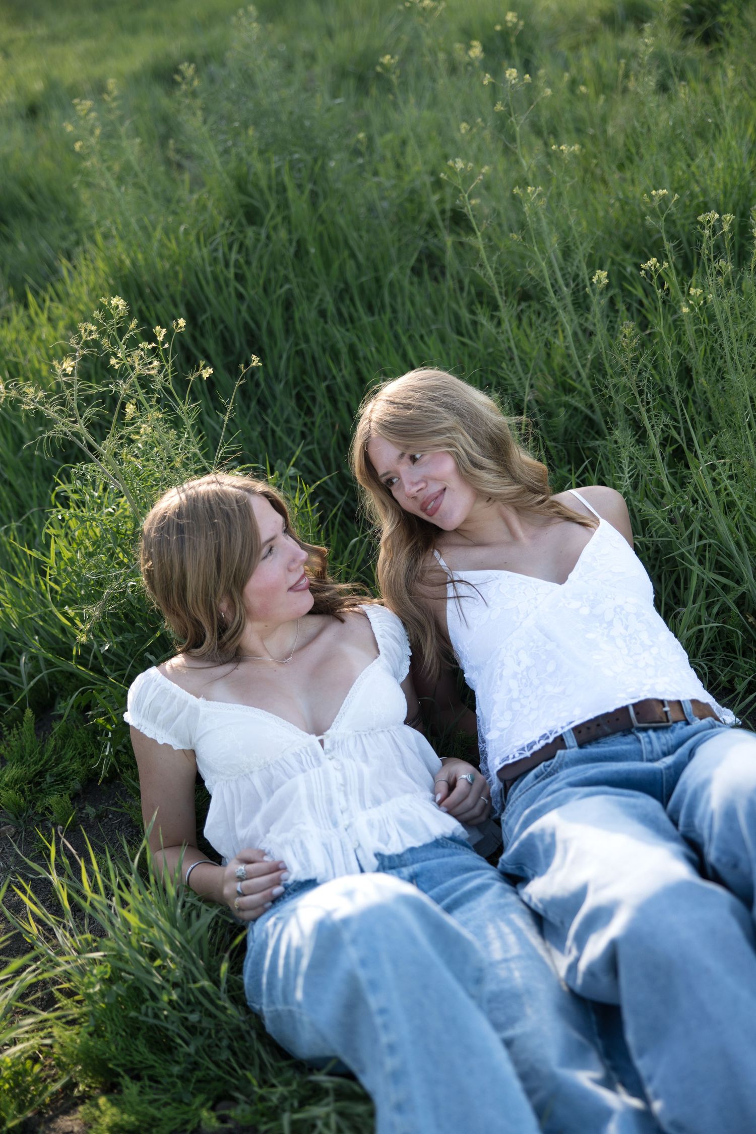 Two young women in white tops and jeans relaxing together in a lush green meadow, smiling and enjoying each other's company.