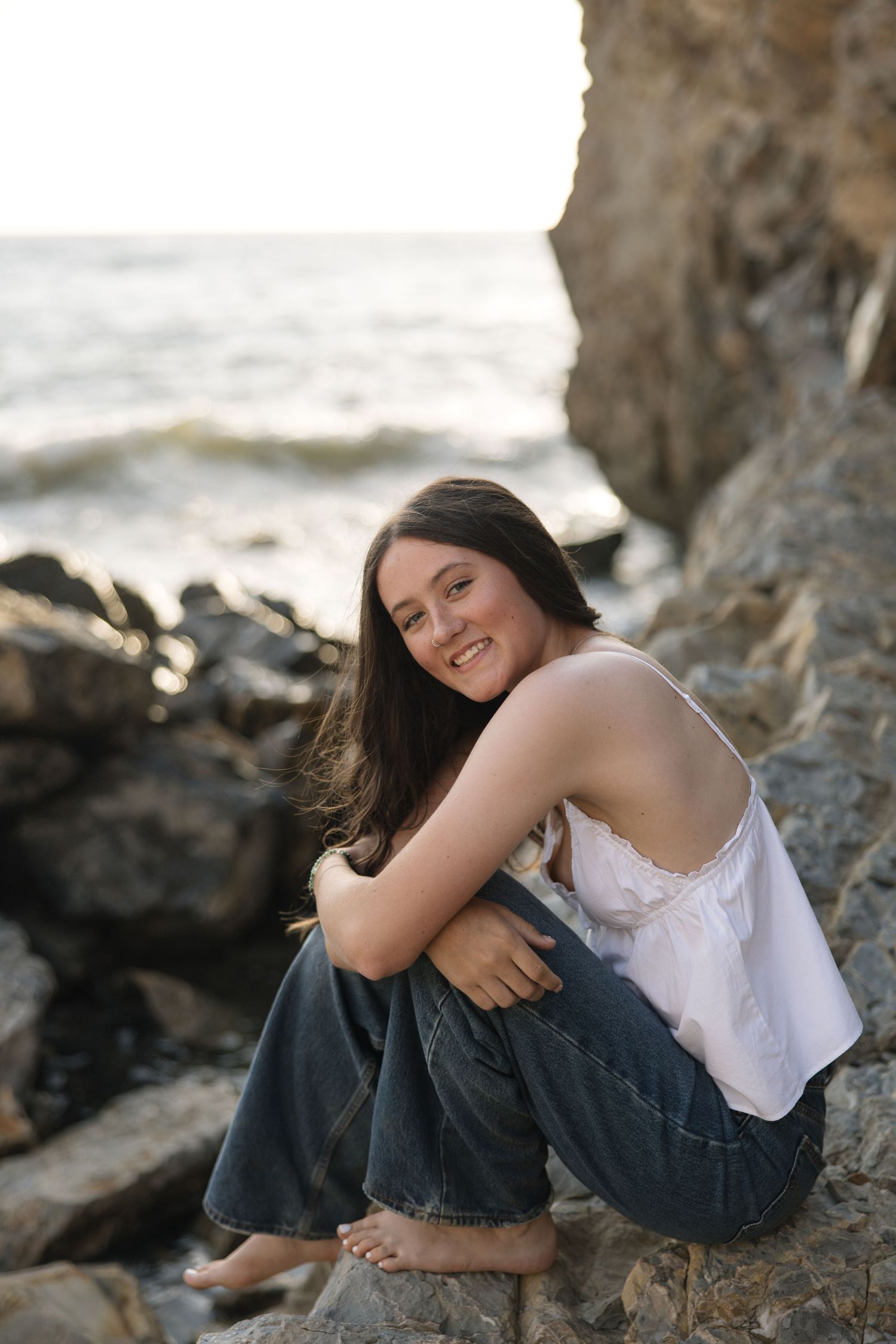 Young woman with long dark hair sits barefoot on rocky beach, smiling warmly in white top and jeans with ocean backdrop.