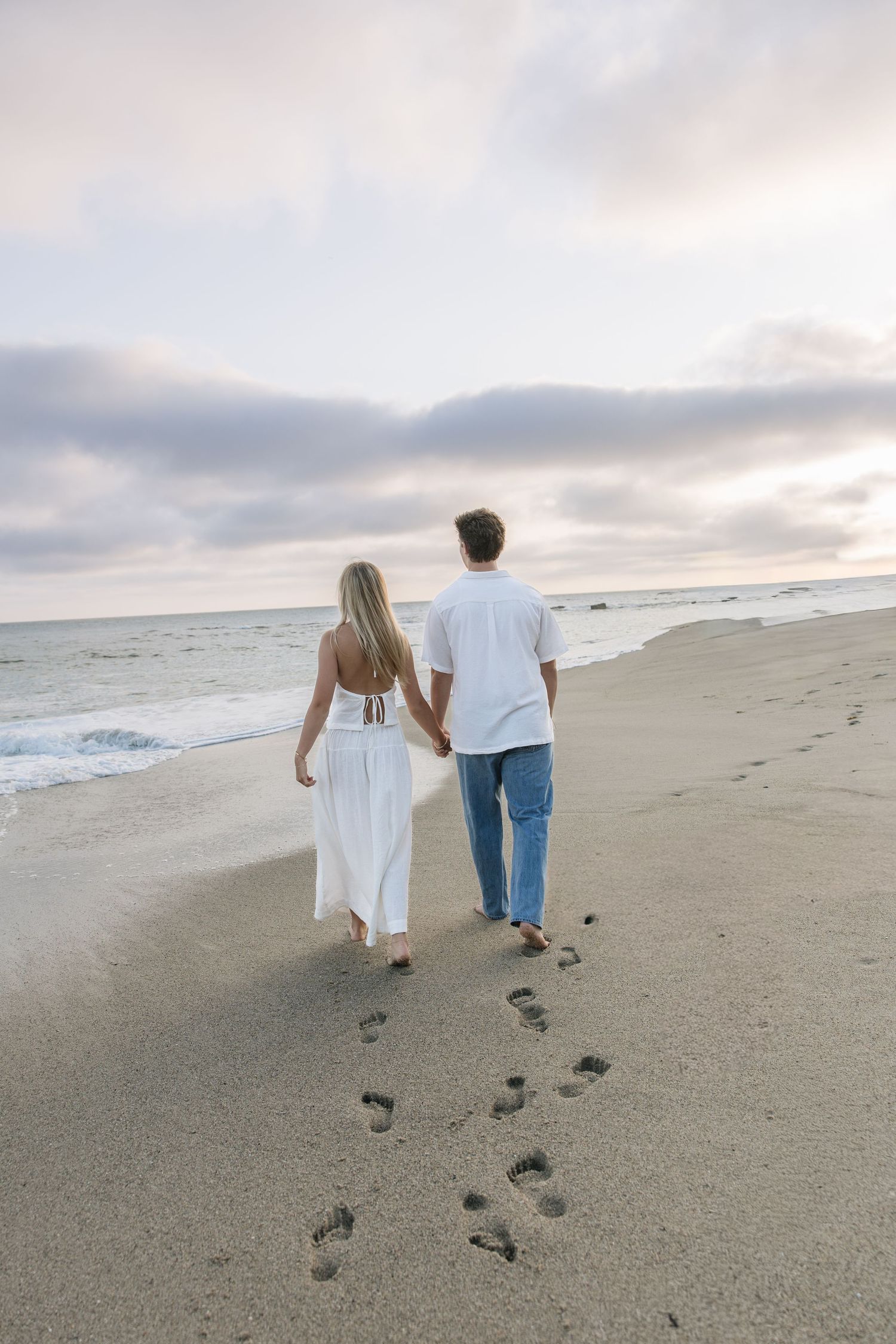 Couple walking hand in hand on sandy beach at sunset, leaving footprints, woman in white dress, man in jeans.