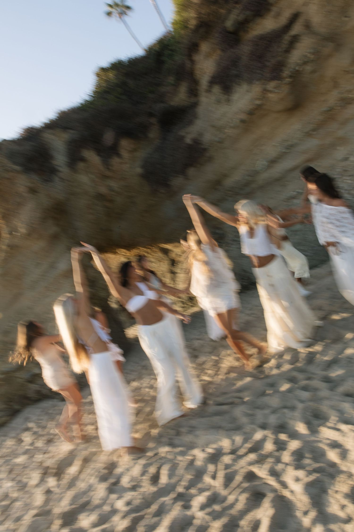 Group of women in white dresses dancing on a sandy beach near rocky cliffs in a motion-blurred photo.