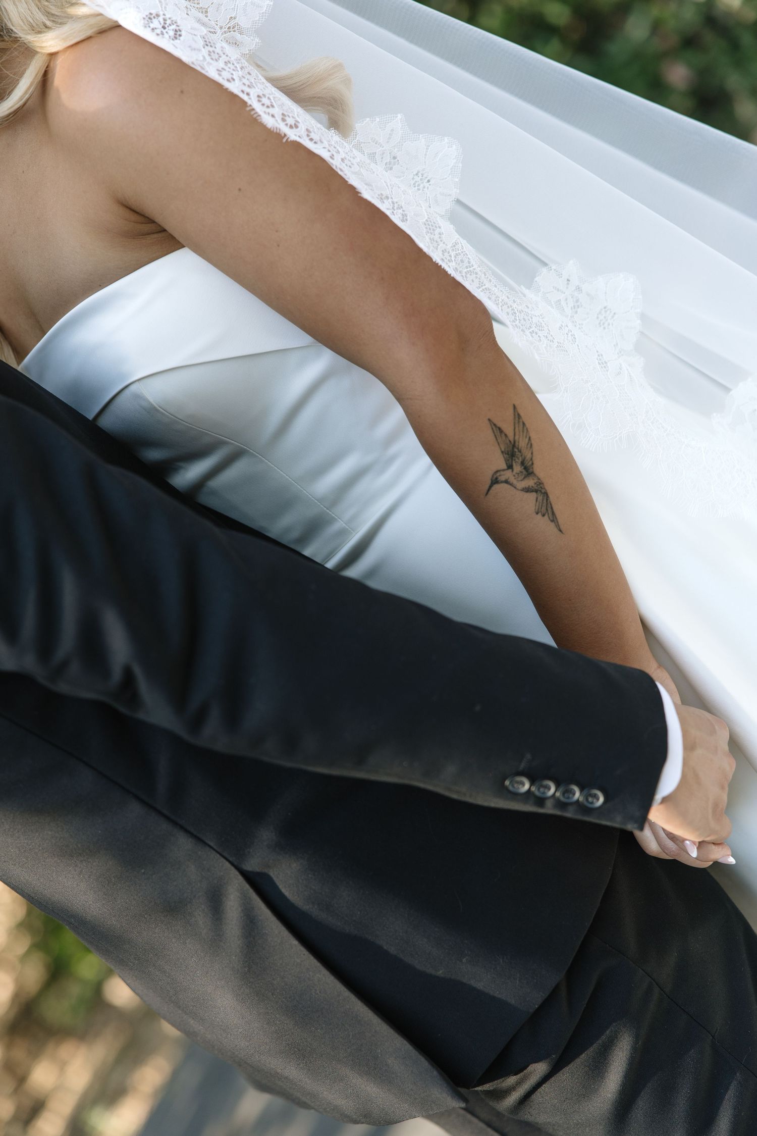 Bride in white lace gown with hummingbird tattoo embracing groom in black suit at wedding ceremony.