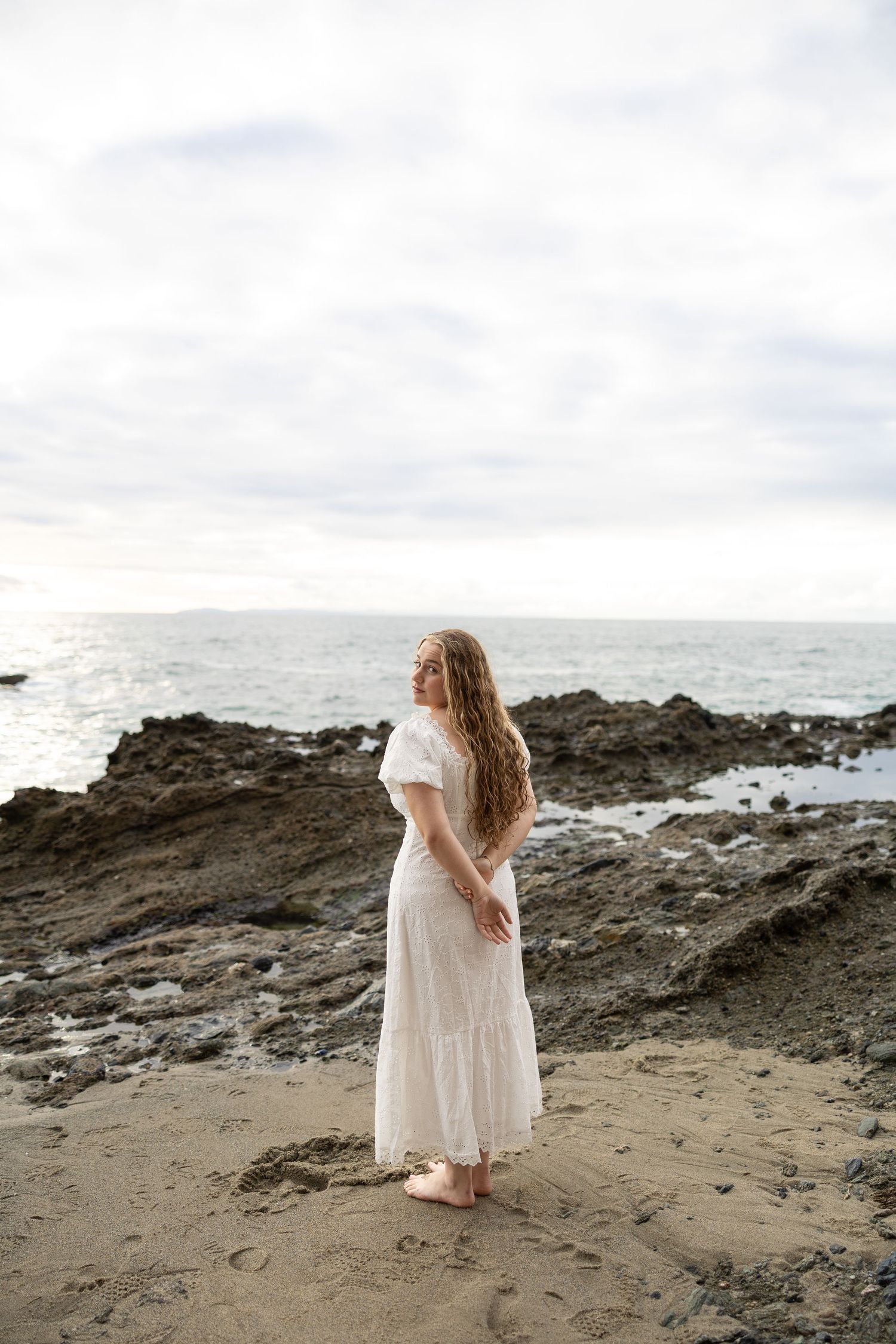 Person in flowing white dress stands on rocky beach coastline at sunset.