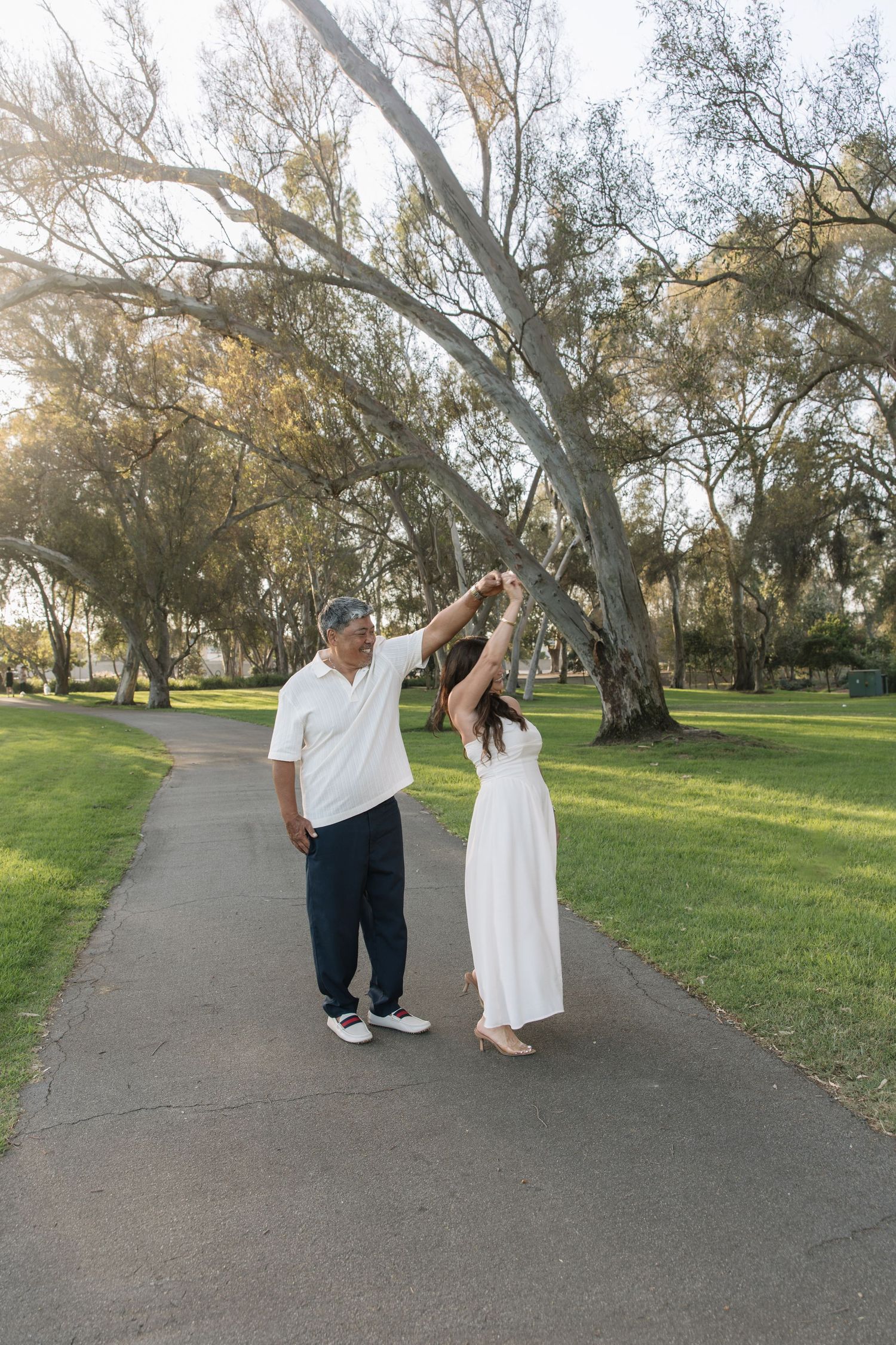 Couple dancing on a paved park path, a man in a white polo twirling a woman in a white dress beneath tall eucalyptus trees.