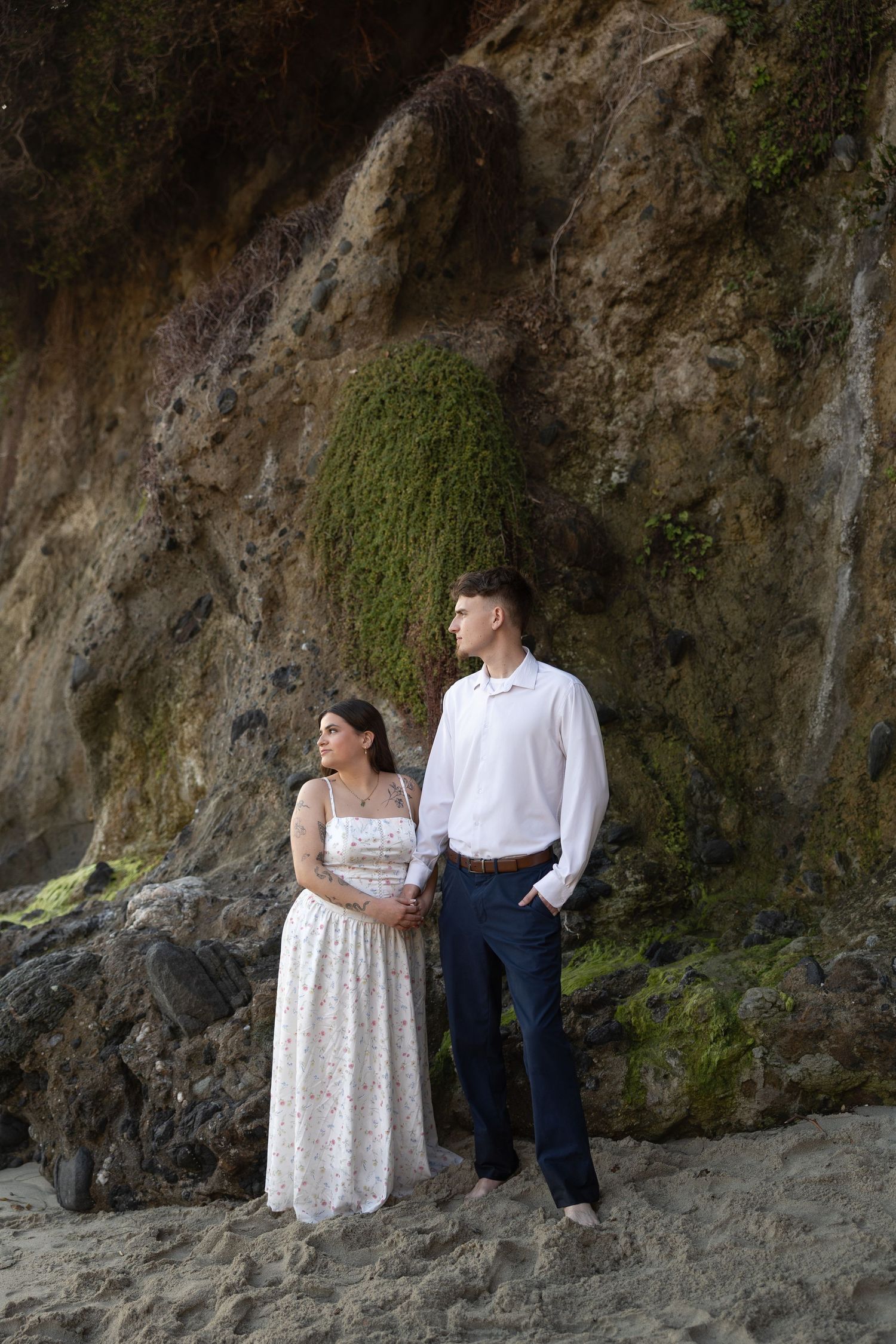 Couple posing against mossy rock wall on beach during portrait session.