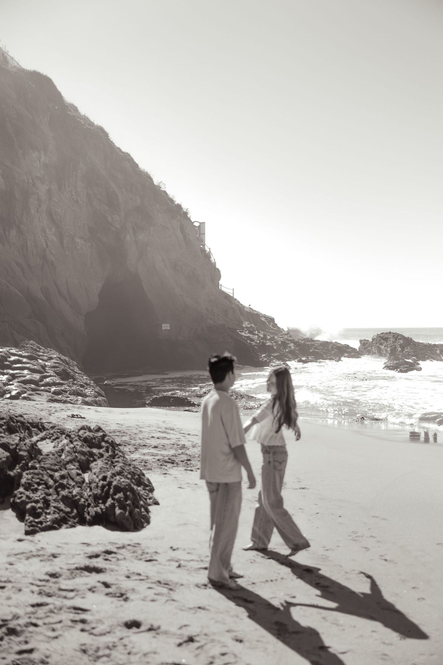 Couple holding hands walking on a sandy beach with rocky cliffs and ocean waves in black and white.