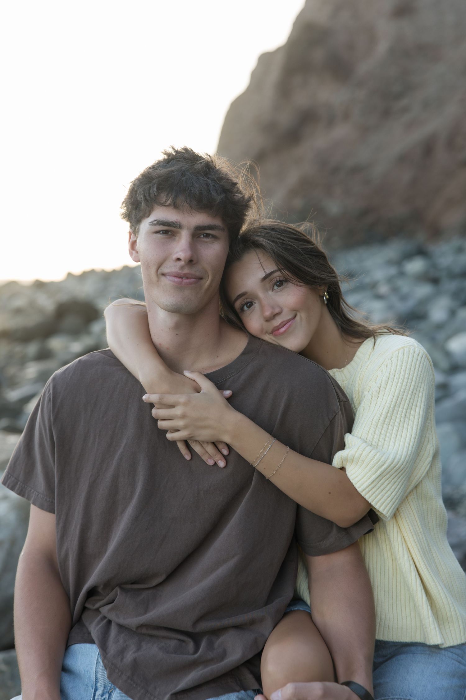 A smiling young woman hugs a young man from behind as they pose together on a rocky beach near a cliff at sunset.