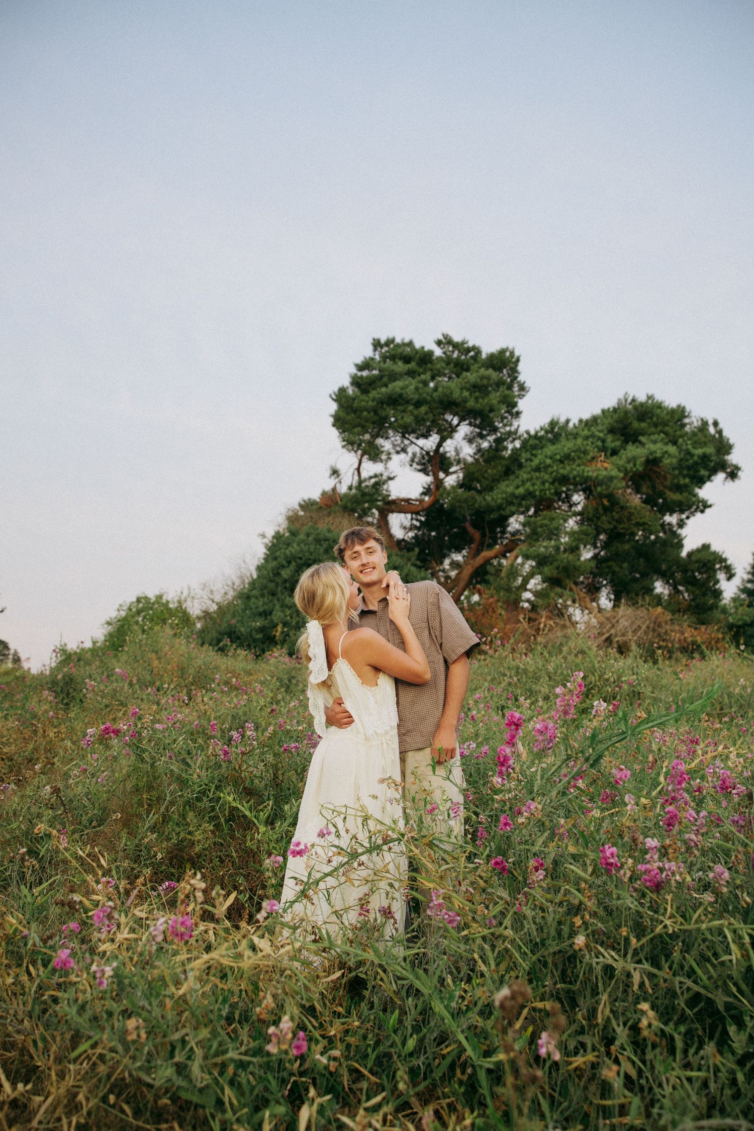 A couple embraces in a wildflower meadow, the smiling man in a tan shirt holds a woman in a white dress outdoors.
