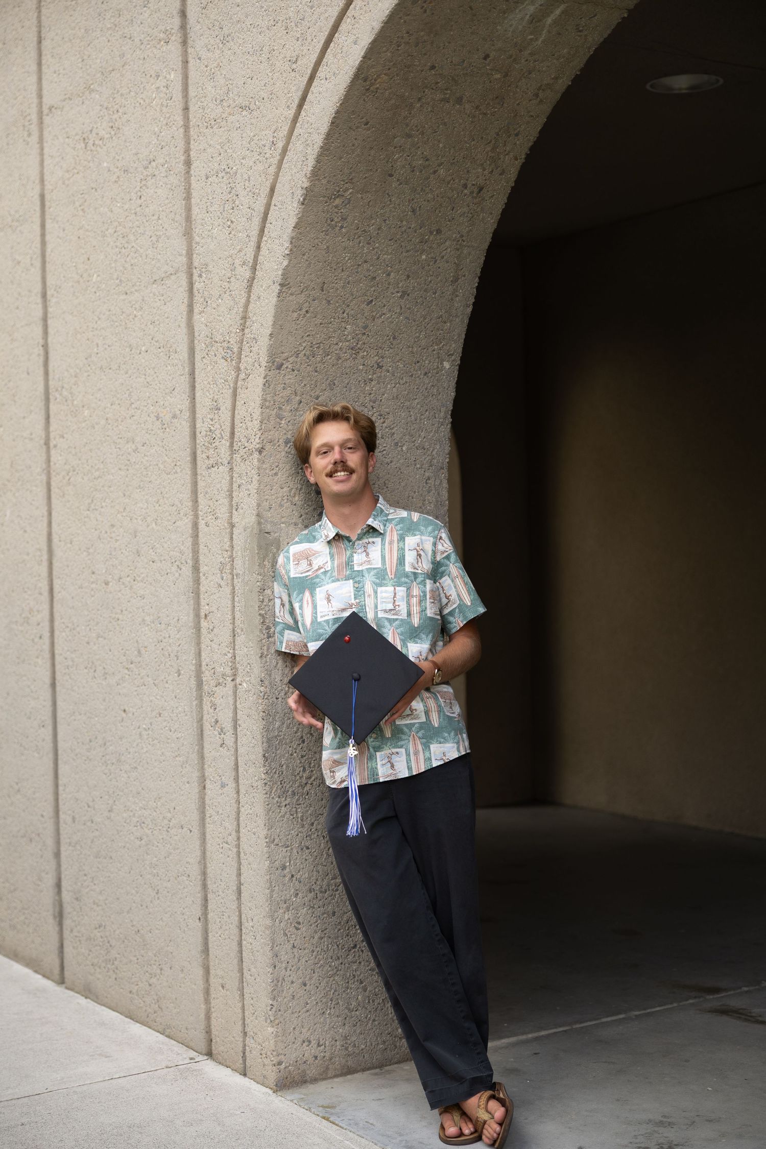 Man in Hawaiian shirt holding graduation cap and diploma, leaning against stone arch at university.