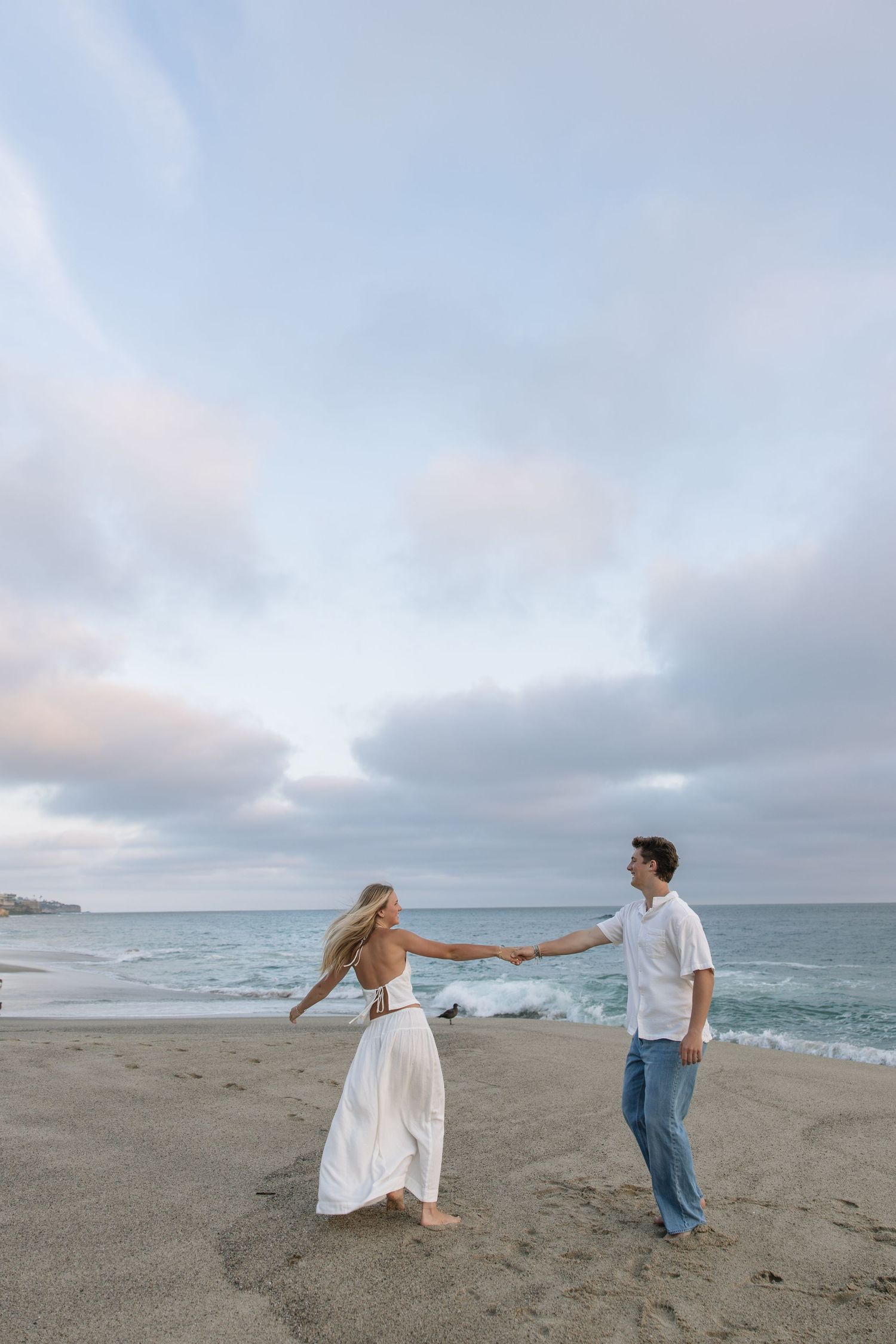 A couple holds hands on a sandy beach at dusk, woman in white dress, man in jeans and white shirt, ocean waves behind.
