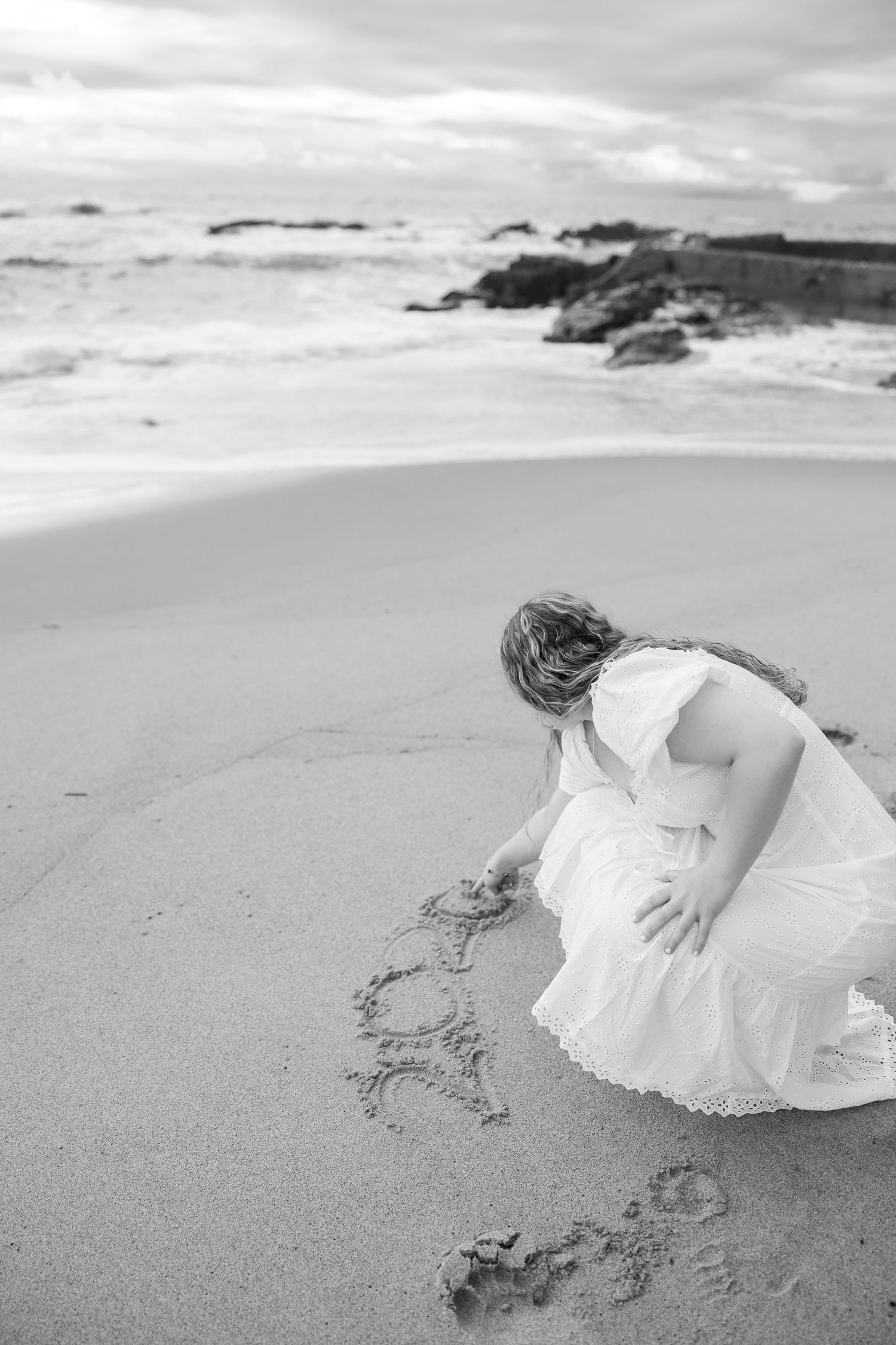 Person in white dress drawing in sand on scenic beach with rocky outcrops.