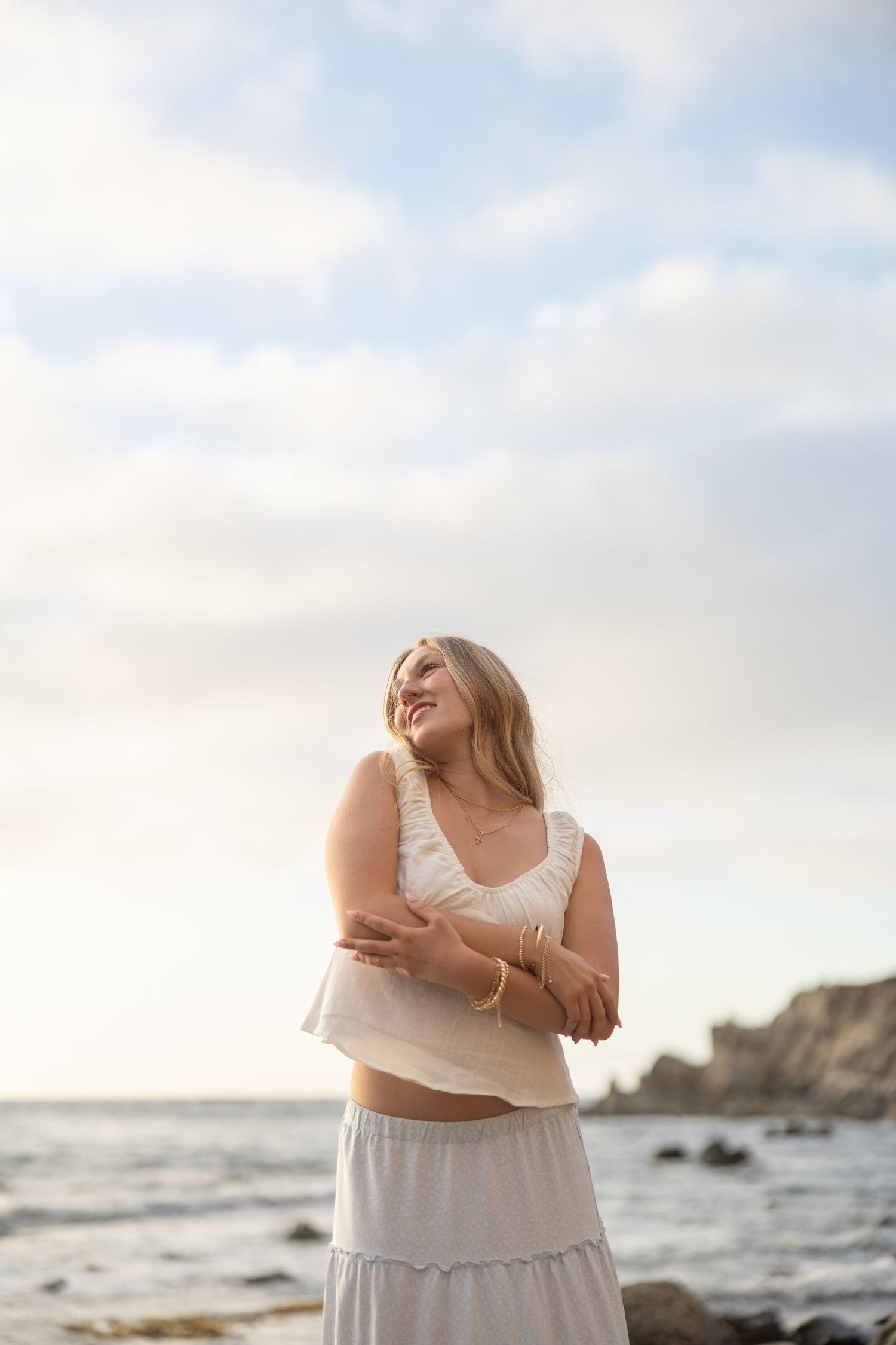 Smiling blonde woman in white crop top and skirt hugging herself on a rocky beach at sunset.
