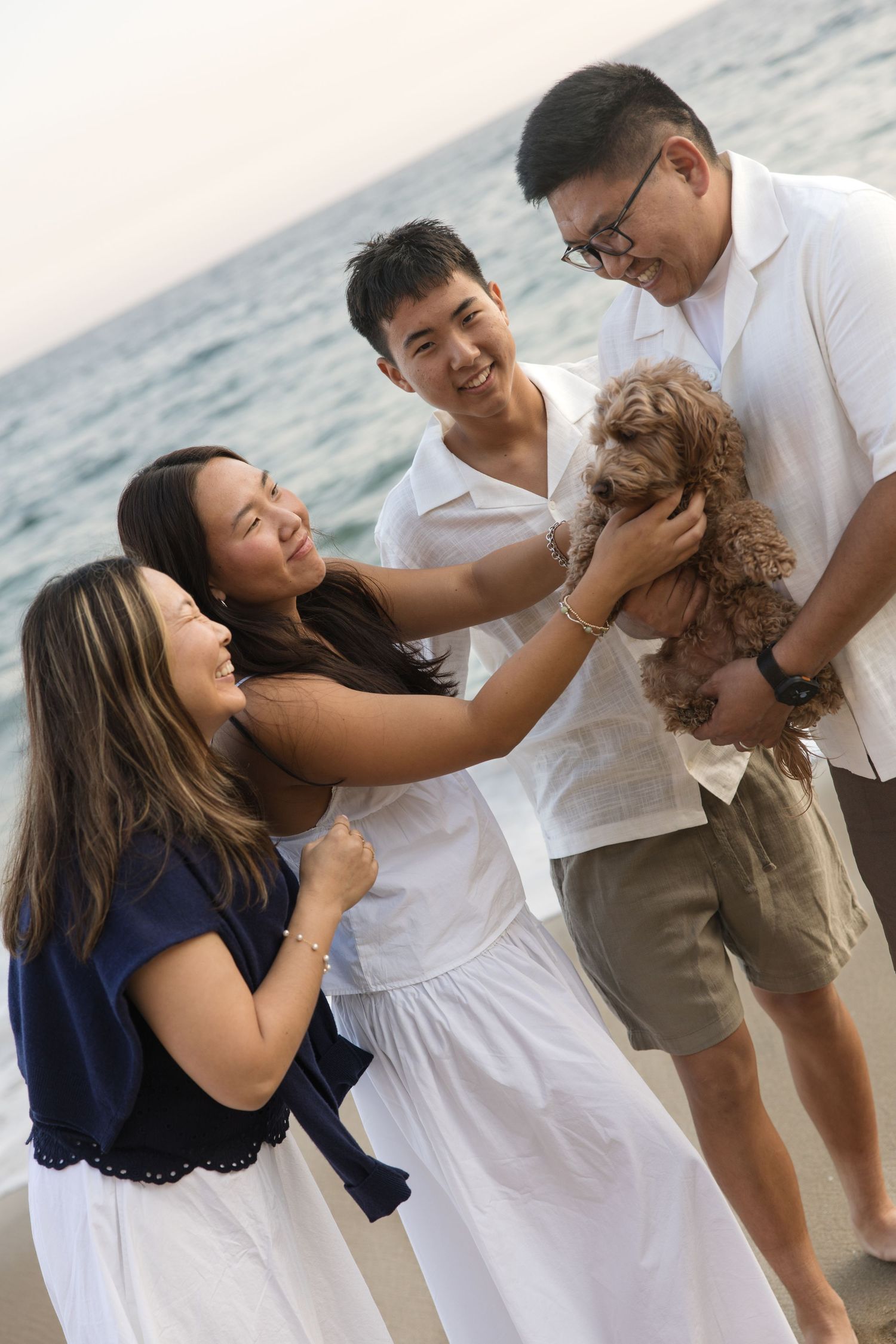 Group of friends smiling and petting a small brown dog on a beach with ocean in the background.