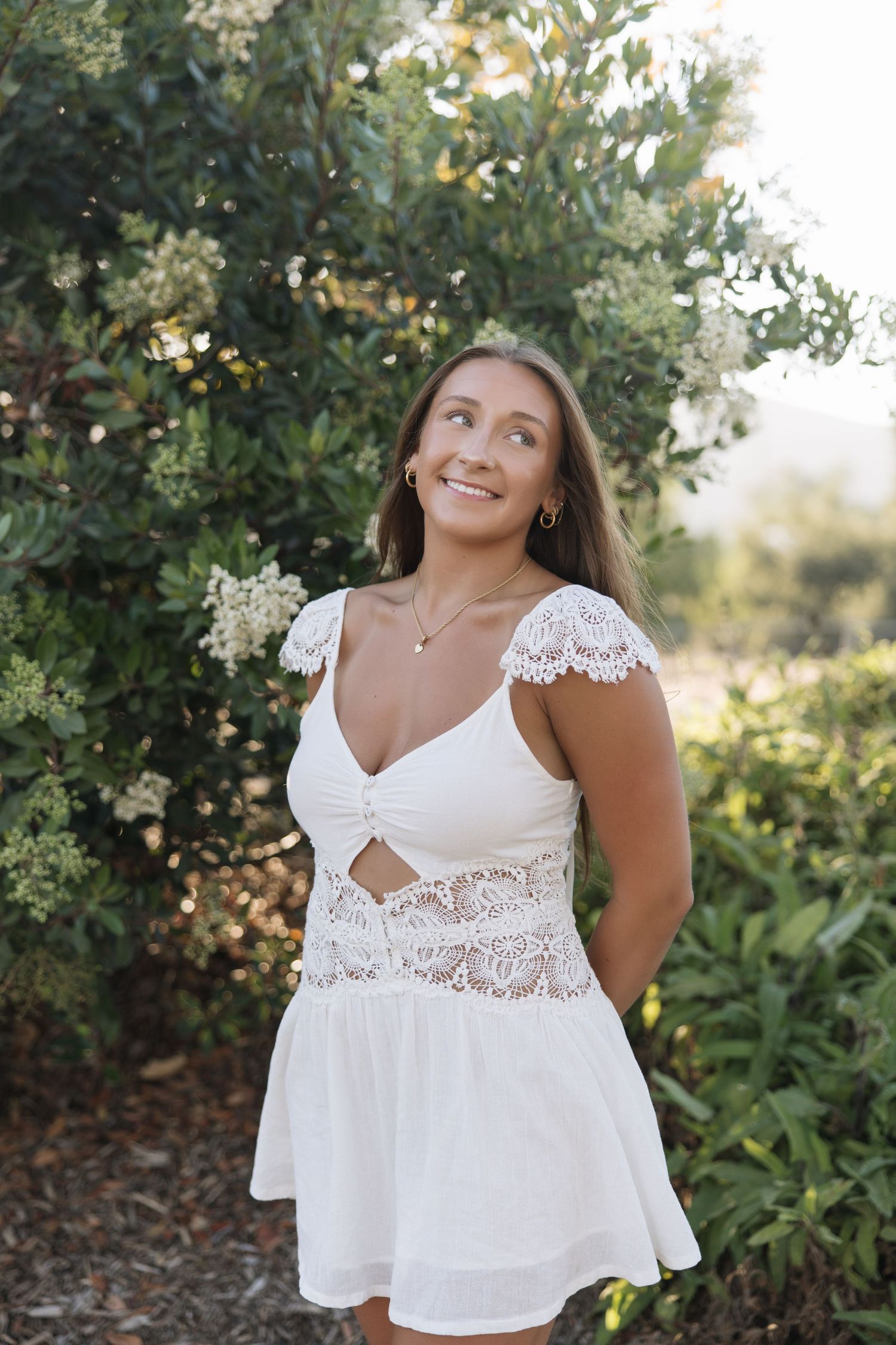 Young woman in white lace mini dress smiling outdoors among green bushes in natural sunlight.