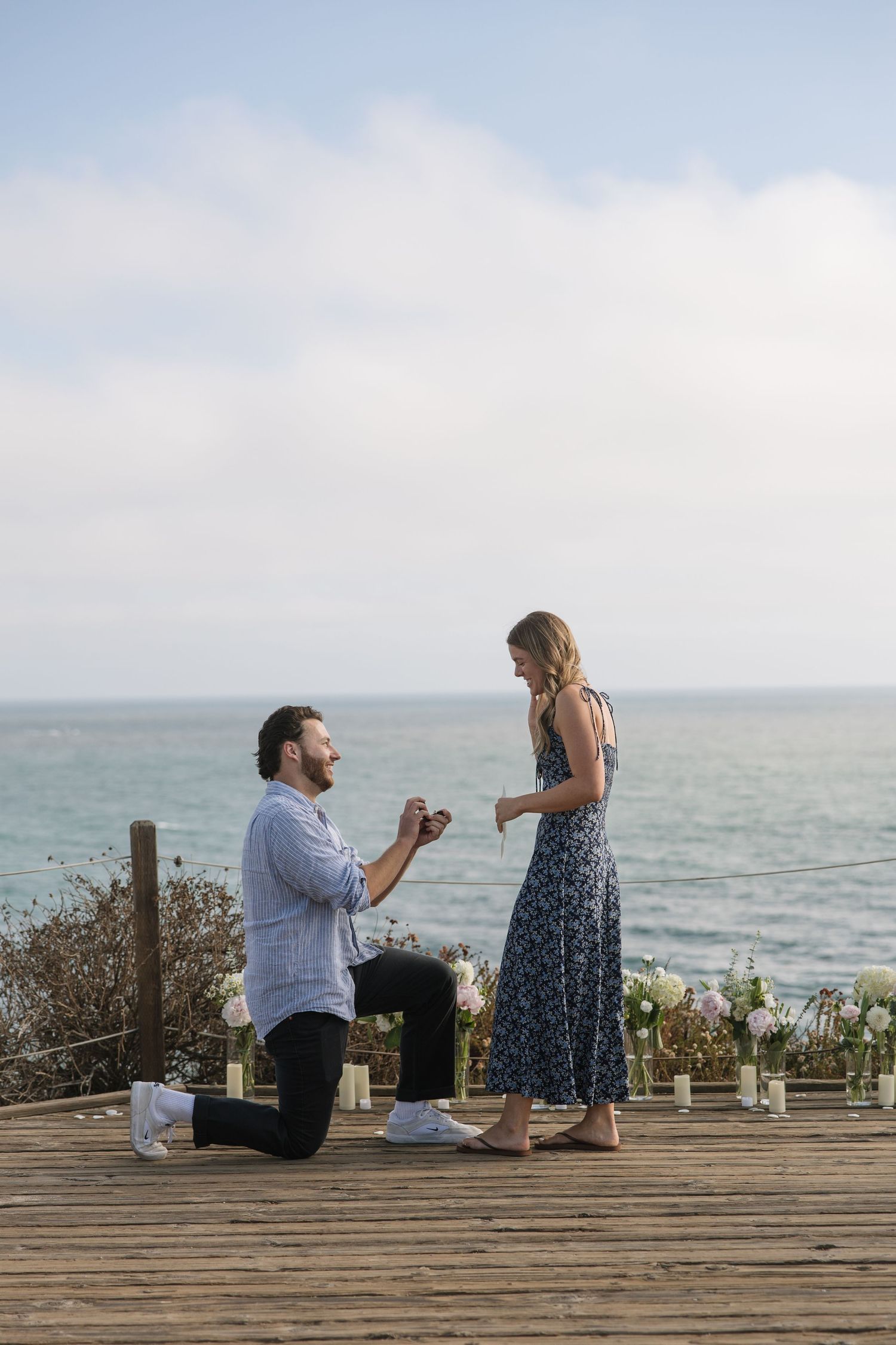 Man proposes on one knee to woman on wooden deck overlooking ocean, with white floral arrangements nearby.