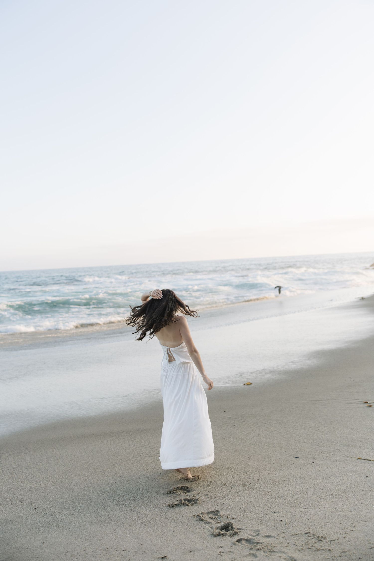 Woman in white dress walking on sandy beach with ocean waves, hair blowing in wind, leaving footprints in sand.
