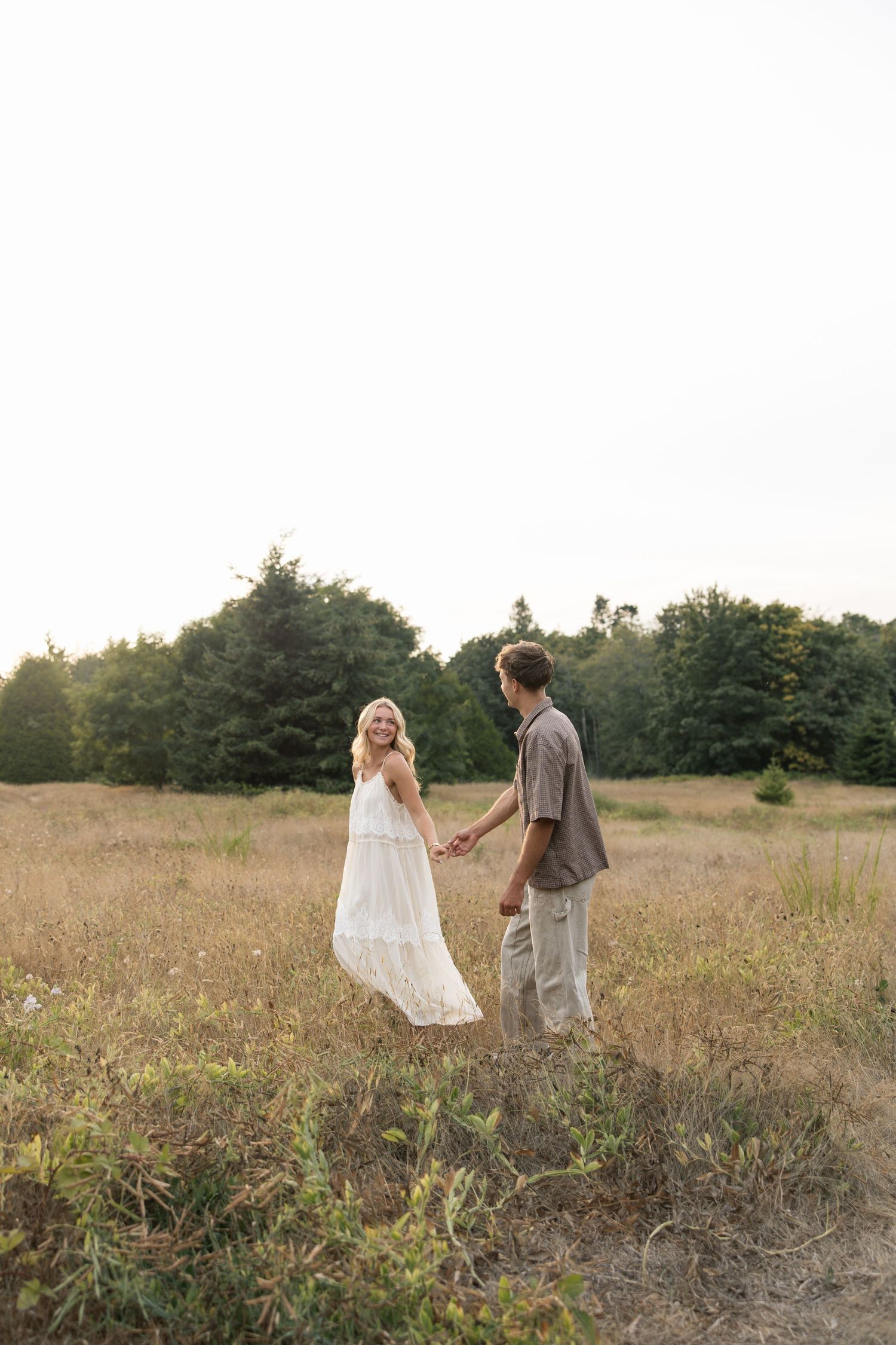 A couple holds hands in a golden meadow, the woman in a white dress smiling back, with green trees in the background.