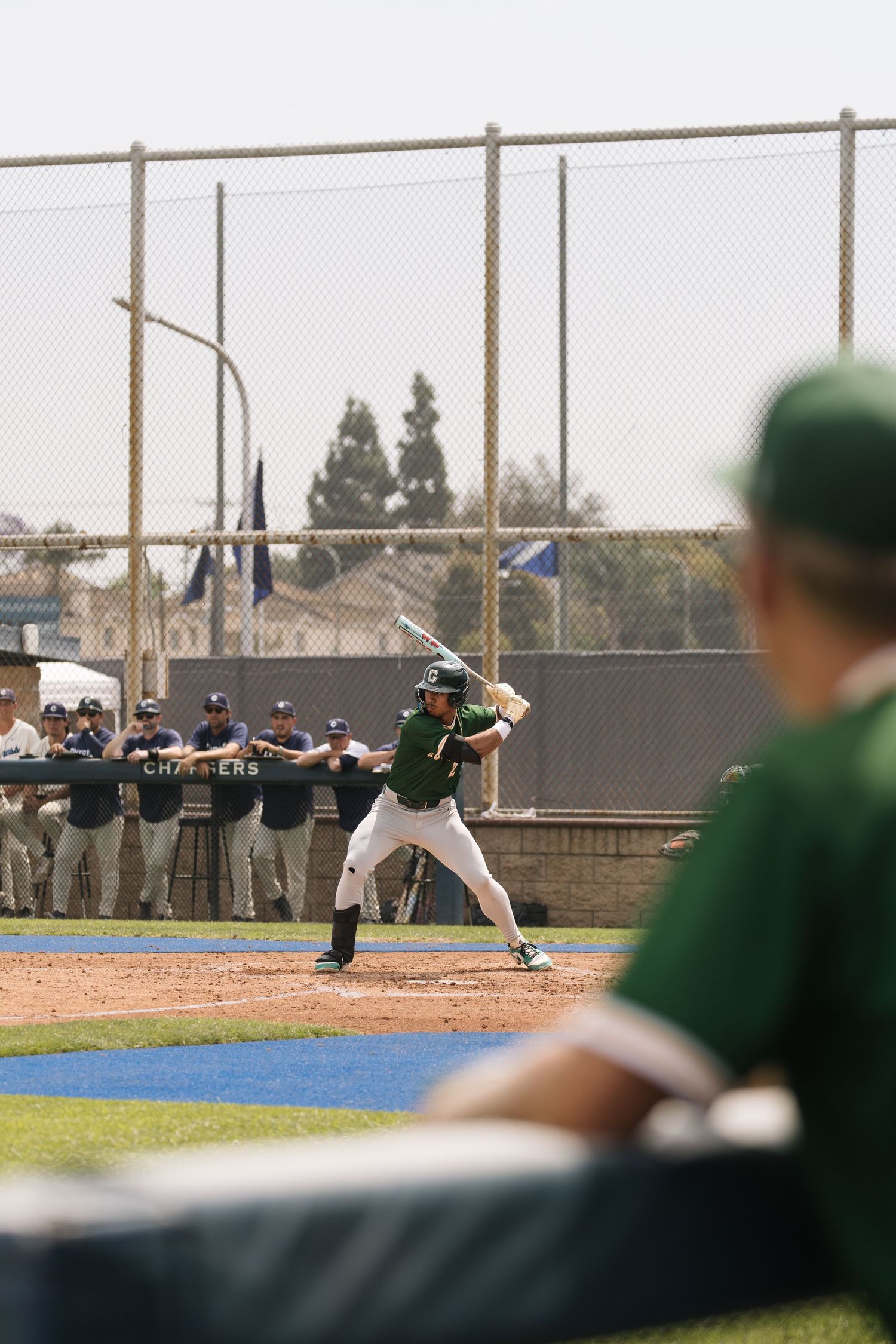 Baseball batter in green uniform at home plate, ready to swing, with dugout and chain-link fence in background.