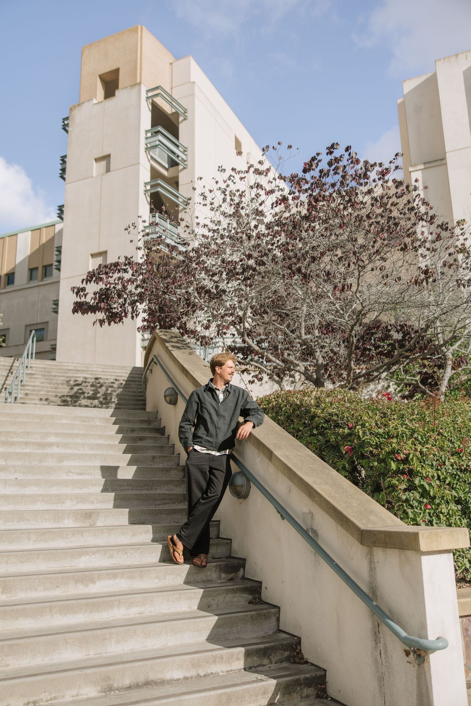 Man in dark jacket leaning on staircase railing outside modern university building with trees and blue sky.