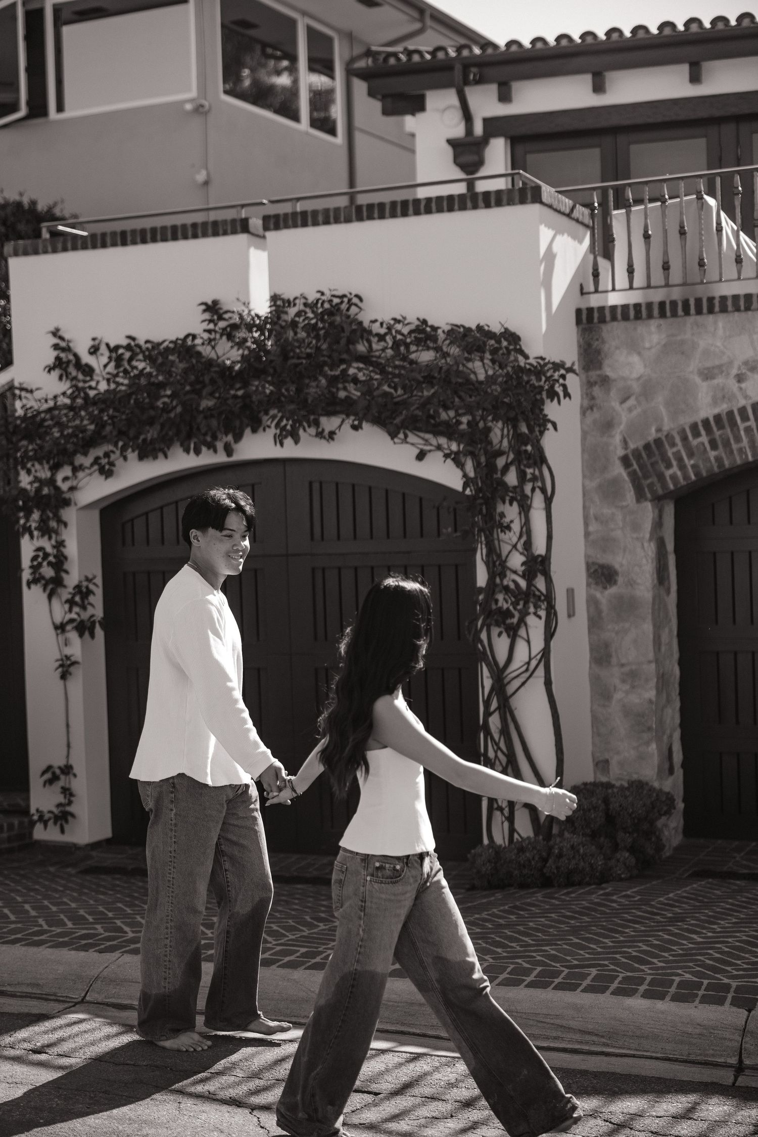 Black and white photo of a young couple holding hands while walking in front of a Mediterranean-style home.