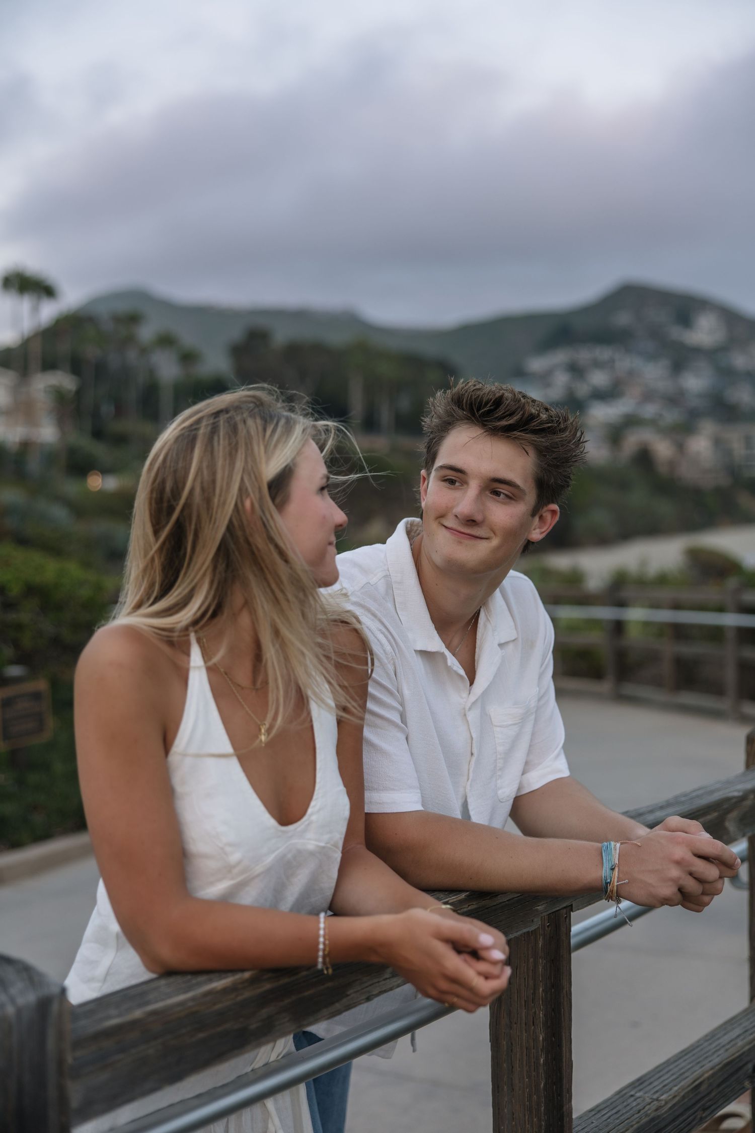 A young couple in white outfits leans on a wooden railing with scenic hills and cloudy sky behind them.